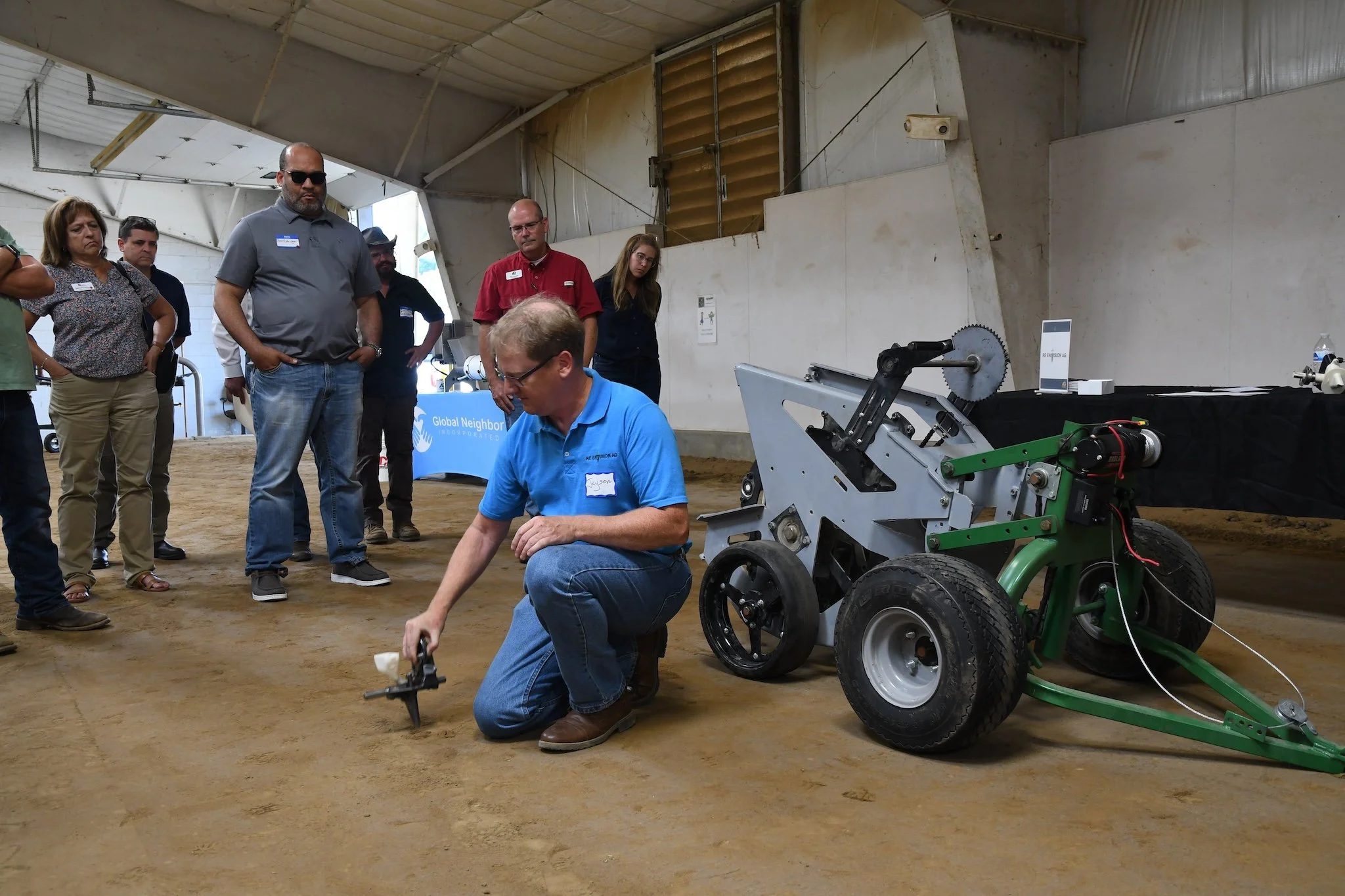 Group of people gathered around a person kneeling and using a tool on a small green and gray robotic vehicle inside a large indoor space with a dirt floor.
