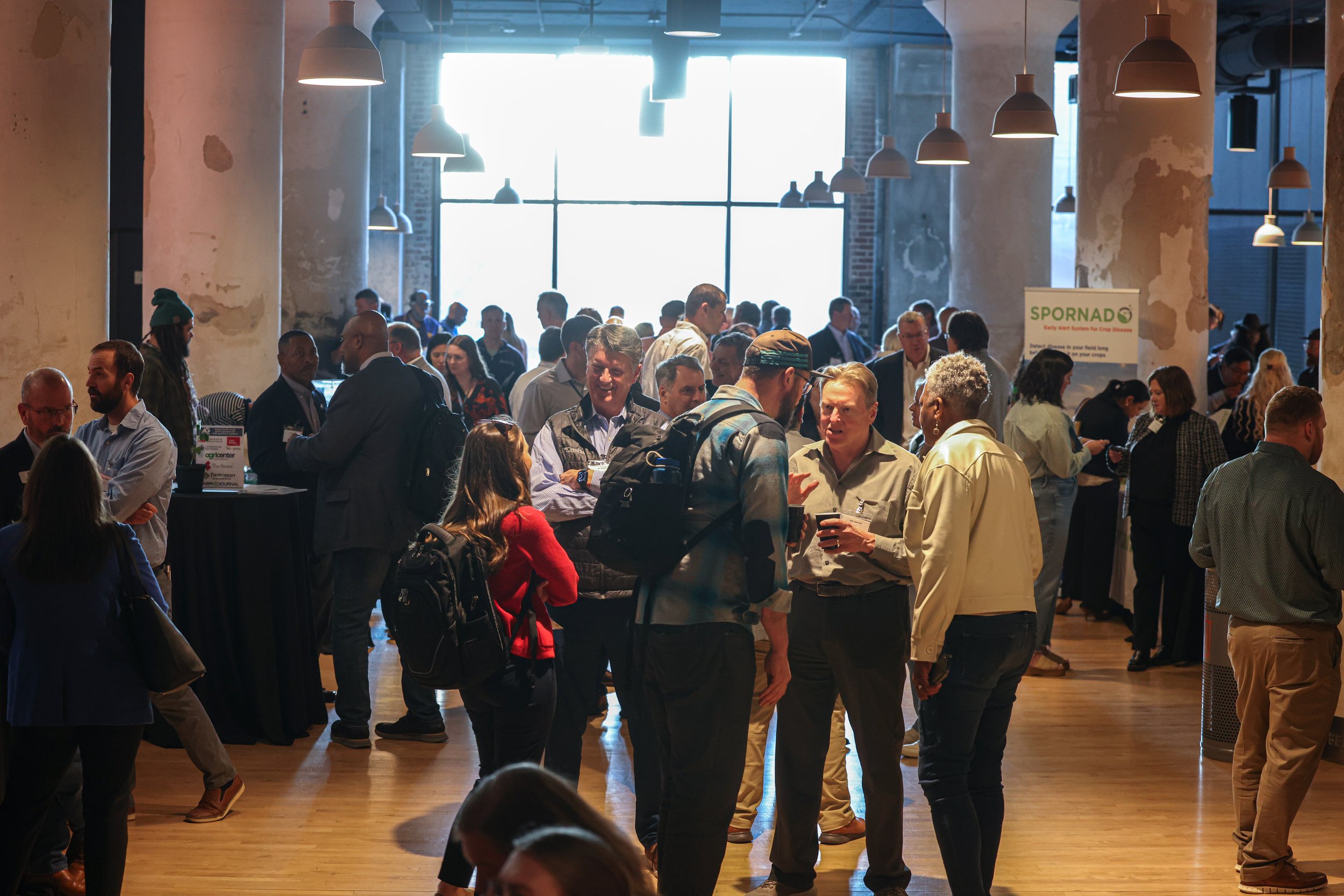 A large indoor conference or networking event with many diverse people engaged in conversations, some holding drinks, in a space with high ceilings, exposed columns, and large windows allowing natural light.