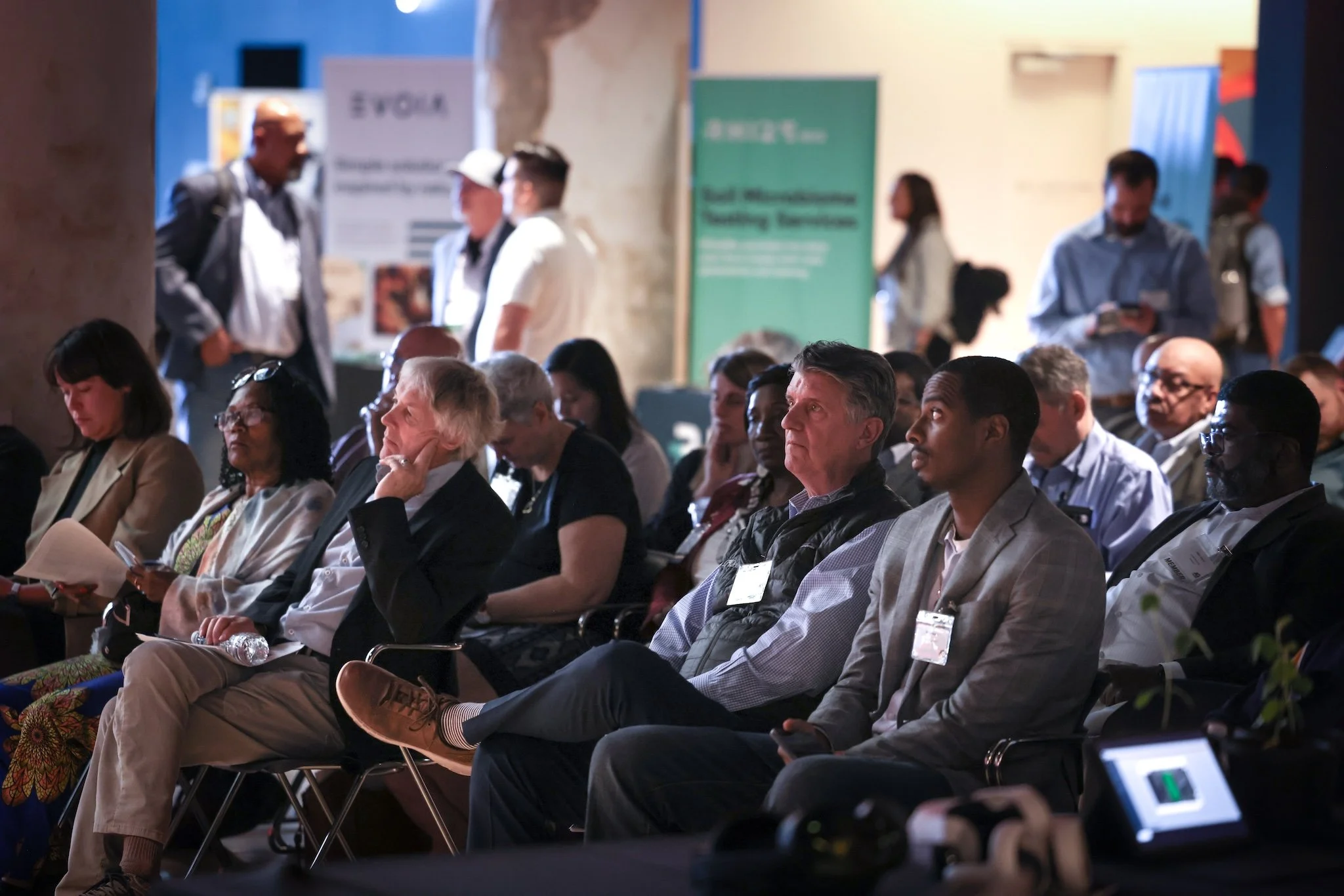 People attending a conference or seminar, seated and listening to a presentation, with some individuals using their phones or taking notes.