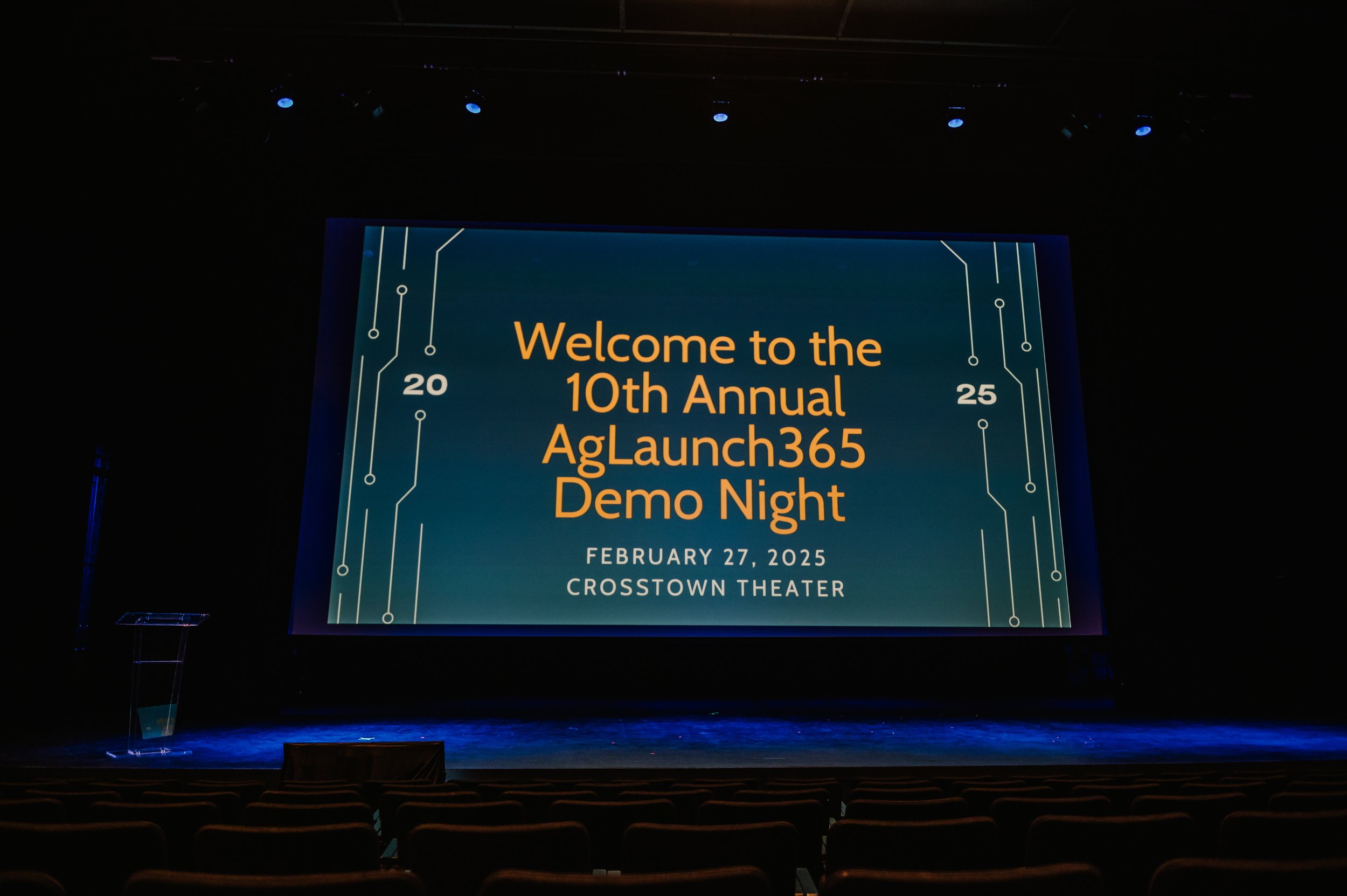 A large digital screen displaying the text 'Welcome to the 10th Annual AgLaunch365 Demo Night' with details 'February 27, 2025, Crosstown Theater'. The screen has a circuit board design border and is set in a dark auditorium with an empty stage and r