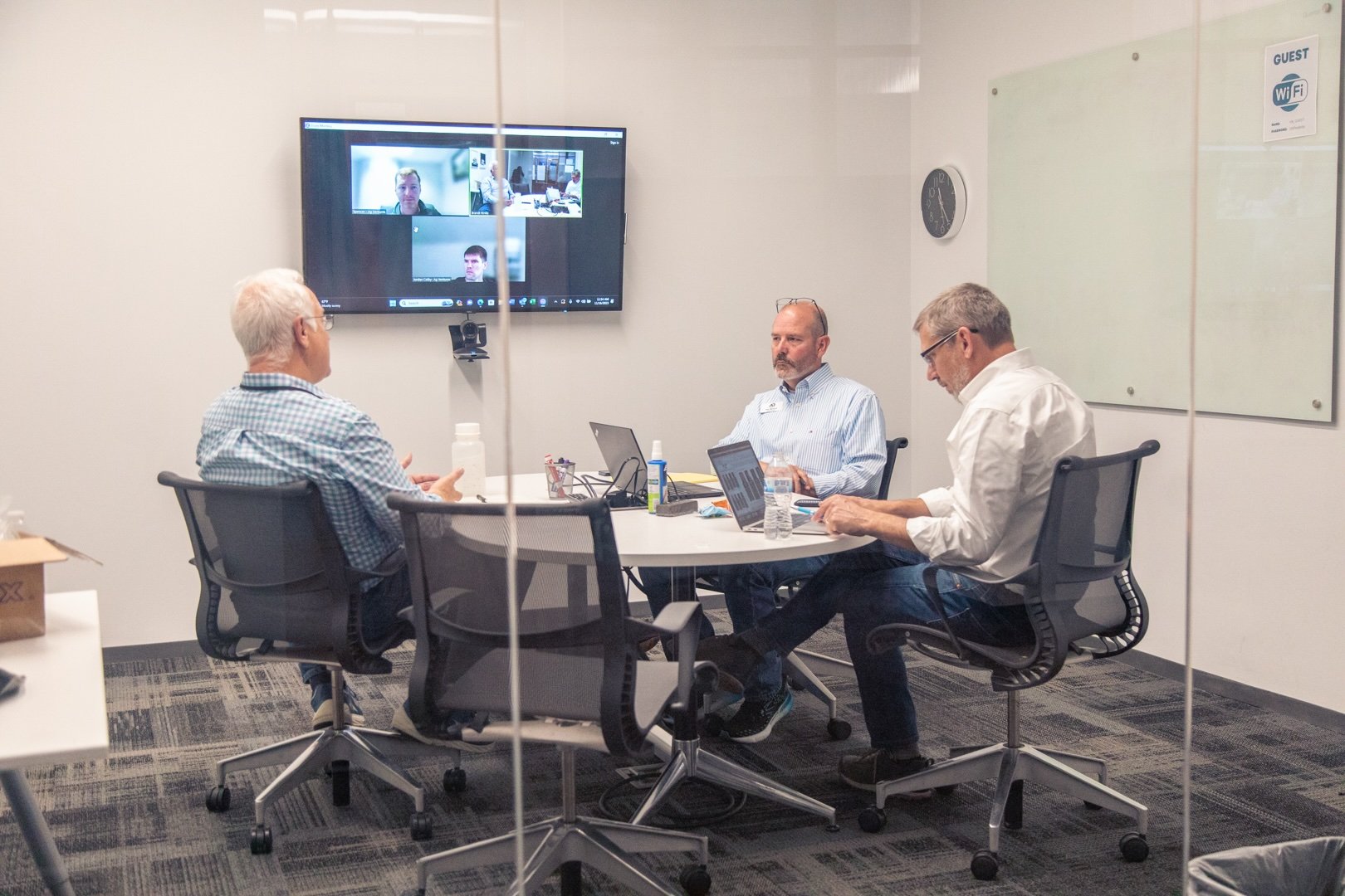 Three men sitting around a table in a conference room, participating in a video conference call displayed on a wall-mounted screen.