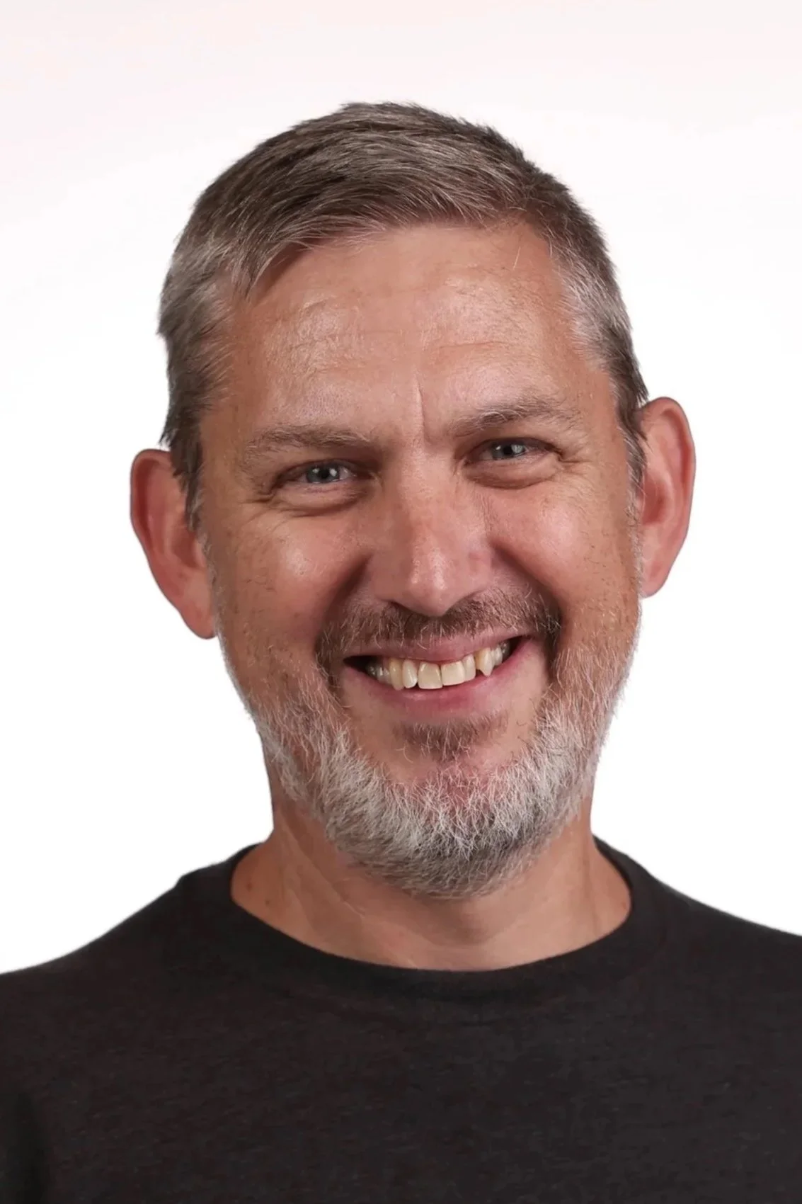 Close-up of a smiling middle-aged man with gray hair and beard, wearing a black shirt against a white background.
