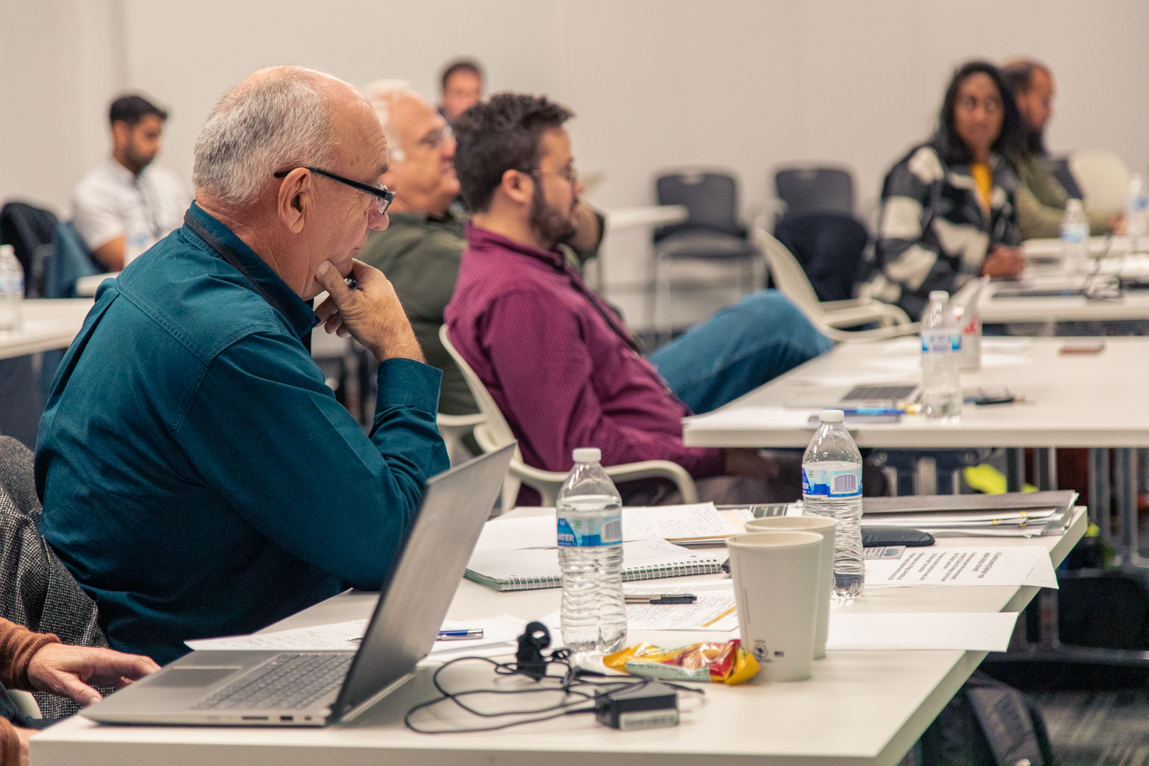 People attending a conference or workshop, sitting at tables with laptops, notebooks, water bottles, and snacks, listening to a presentation in a conference room.