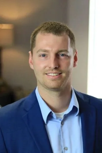 A young man with short brown hair, wearing a blue suit jacket and a light blue shirt, smiling in an indoor setting with a window and lamp in the background.