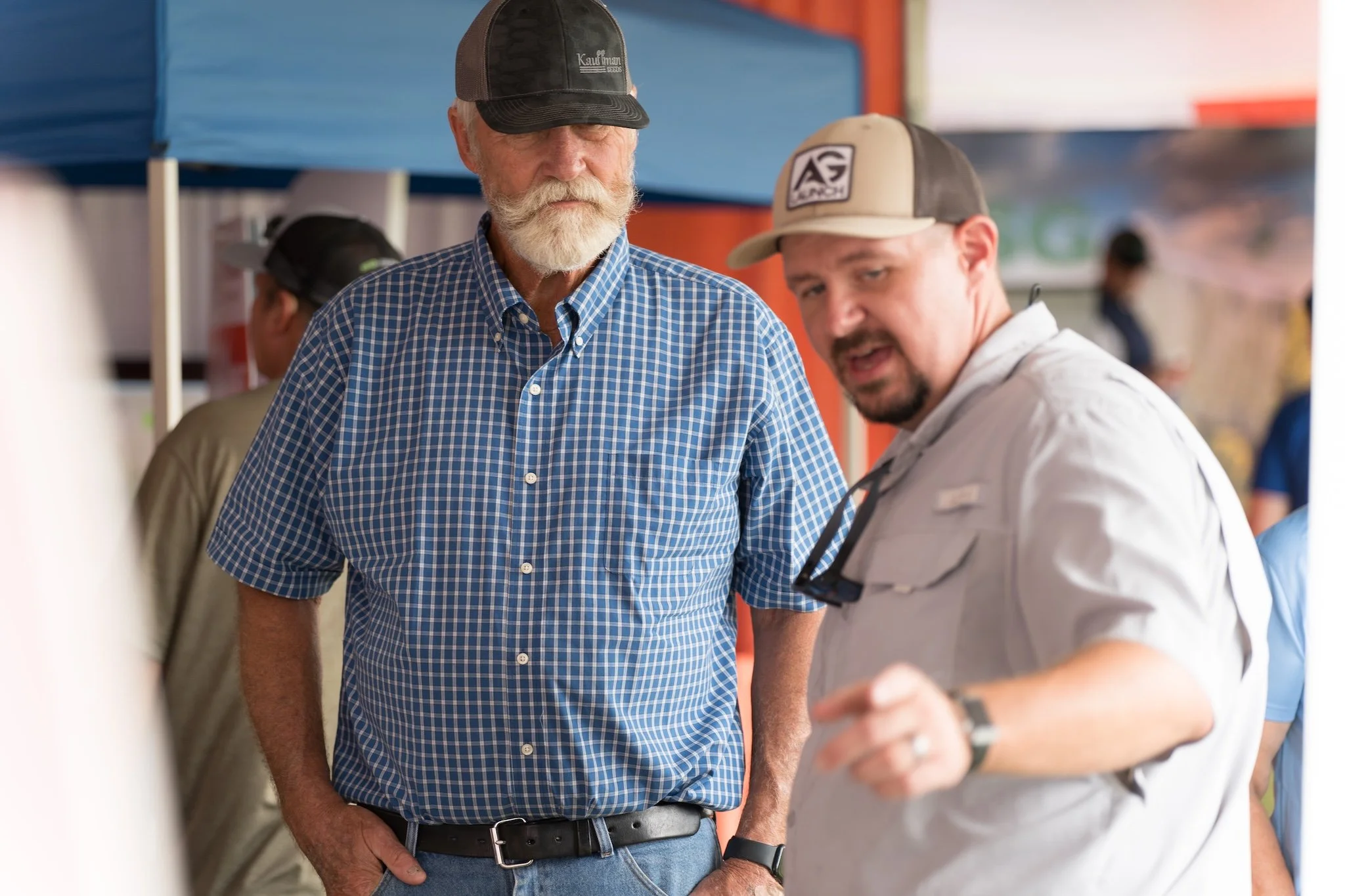 An older man with a white beard wearing a blue checkered shirt and a black cap stands next to a man with a mustache and a goatee wearing a light-colored cap and a gray shirt. They appear to be in a busy outdoor setting, possibly a market or fair, wit