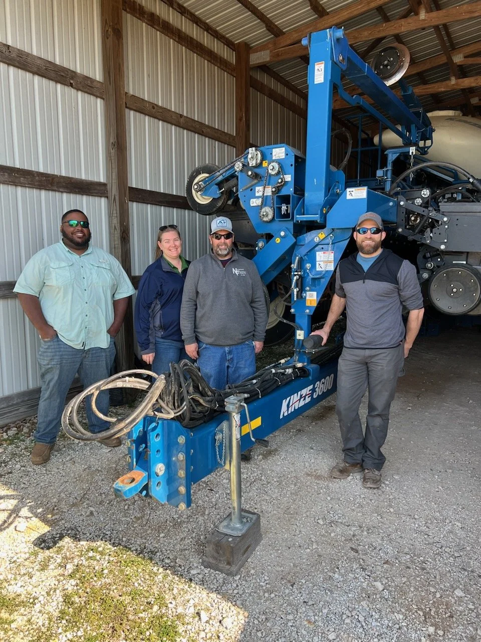 Four people standing next to a large blue industrial machine in a barn or warehouse with metal walls and exposed wooden beams.