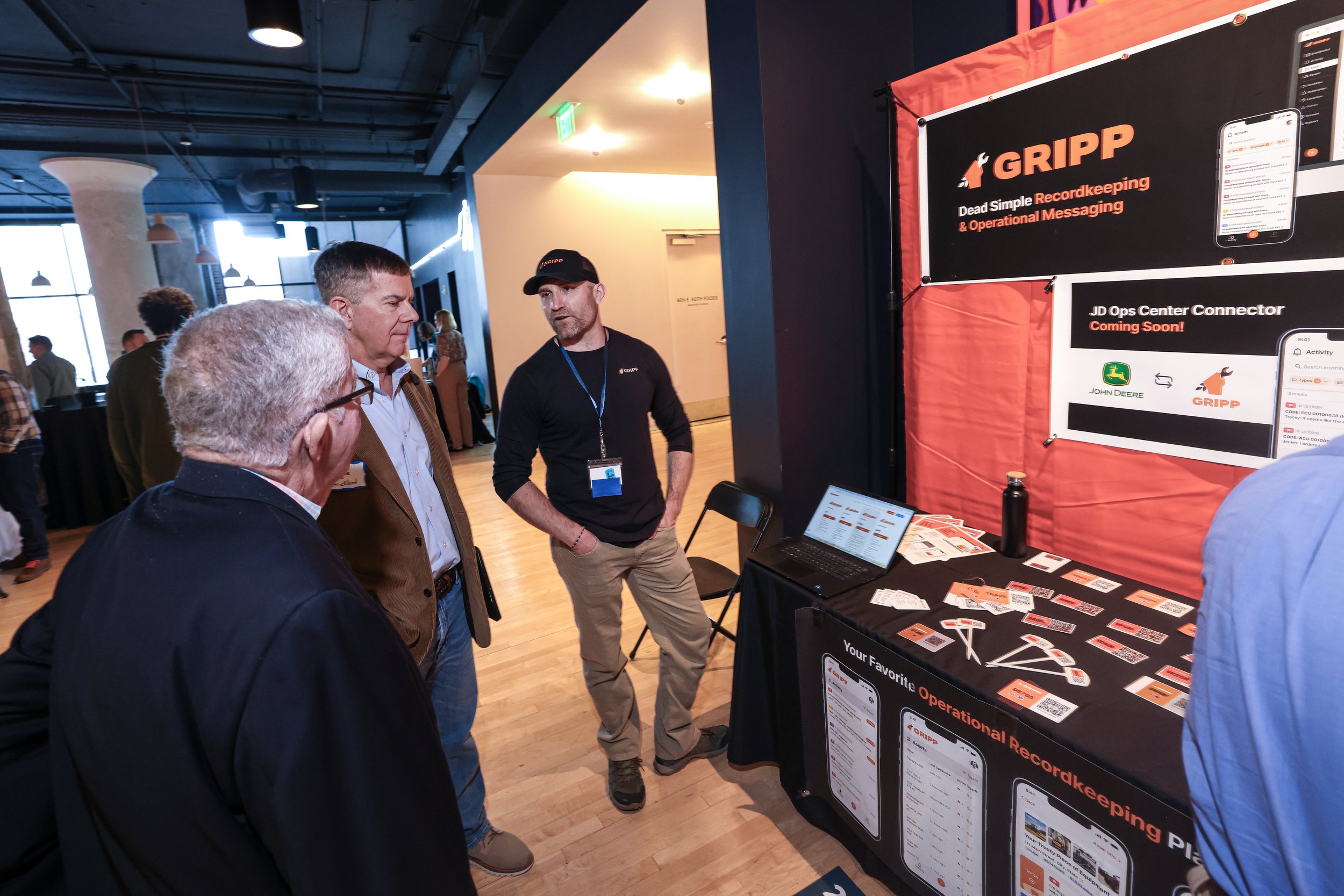 People attending a booth at a conference promoting GRIPP, a tool for recordkeeping and operational messaging. Two men are engaged in conversation with a representative by the display table.