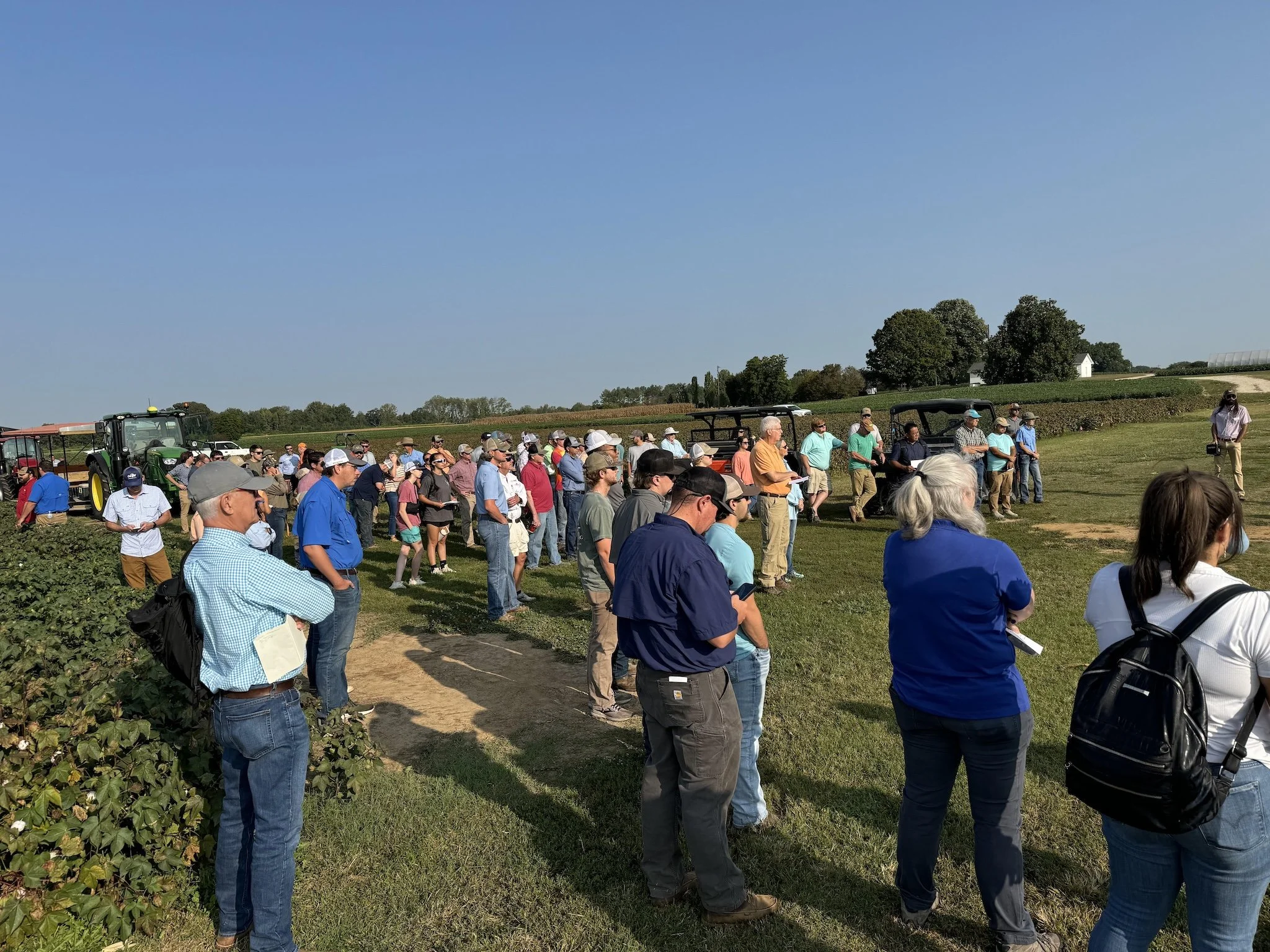 A group of people gathered outdoors on a farm, some standing near tractors and utility vehicles, with fields, trees, and a clear blue sky in the background.