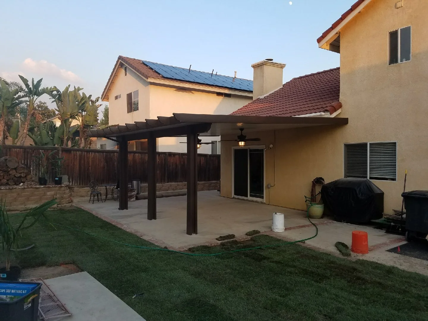Backyard patio with a concrete slab, a wooden pergola, potted plants, a barbecue grill covered with a black cover, and two orange and white buckets near the house’s beige wall with a sliding glass door. The yard has a lawn and a wooden fence with nei