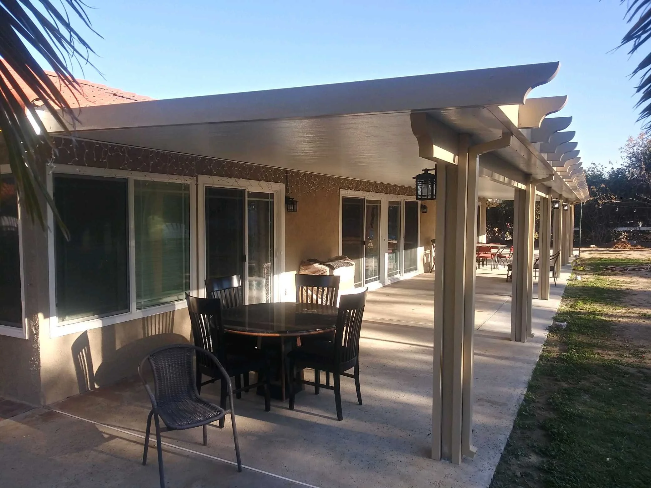 Patio with black chairs and a round table, covered by a pergola, attached to a beige house with sliding glass doors, in a sunny outdoor setting.