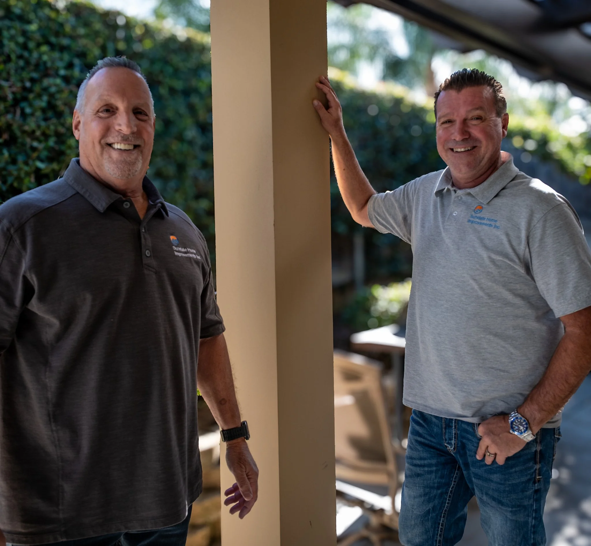 Two smiling men standing outdoors, one resting hand on a beige column, both wearing gray polo shirts with a company logo.