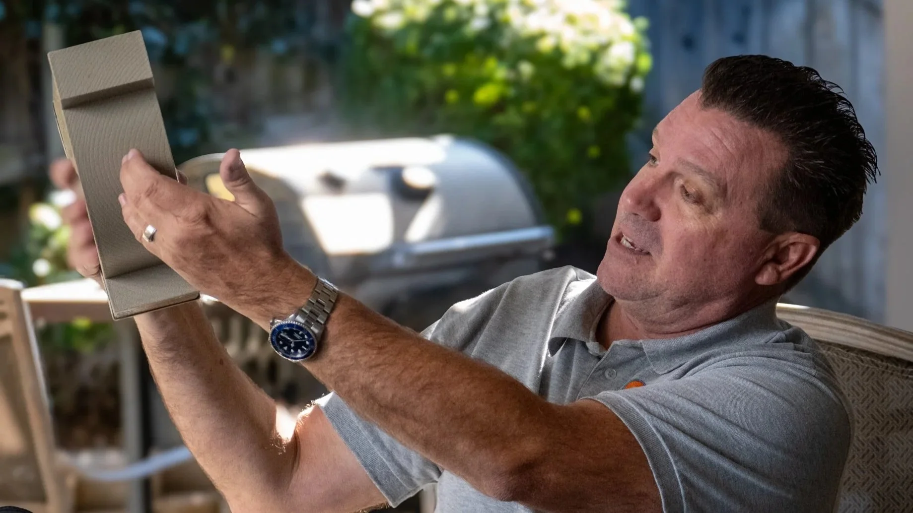 A man sitting outdoors in a backyard, holding a tablet device and looking at it with a focused expression.