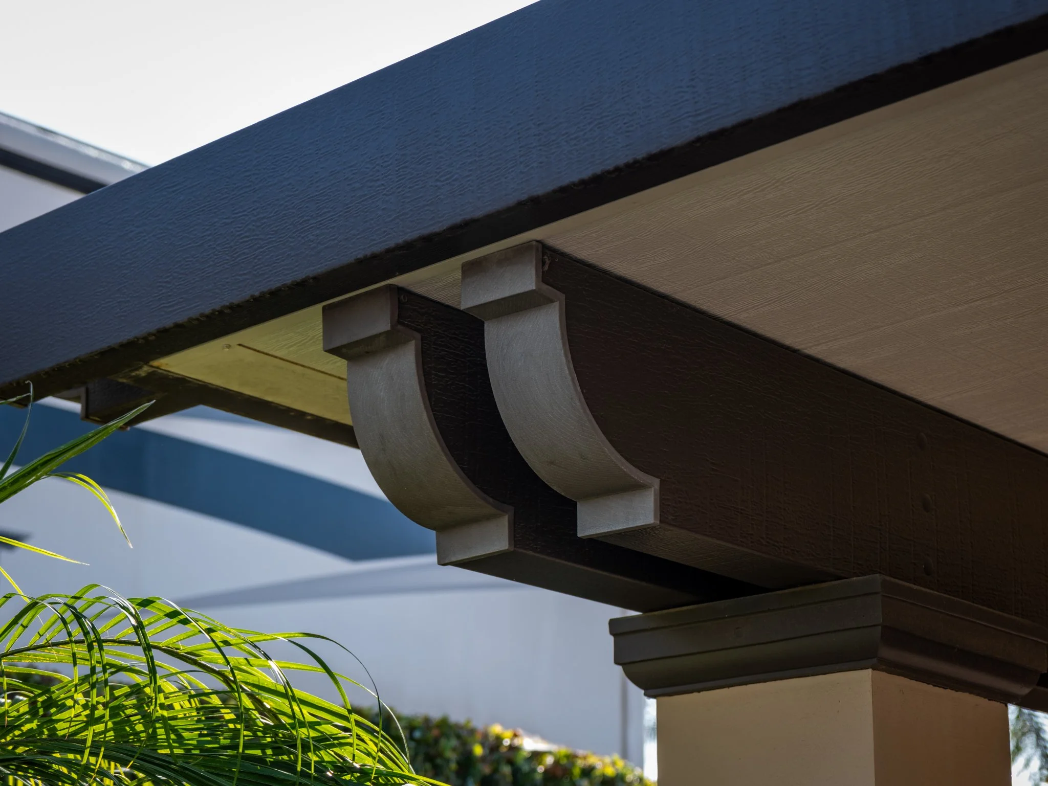 Close-up of a modern architectural overhang with decorative brackets, part of a building structure, with some greenery in the background.