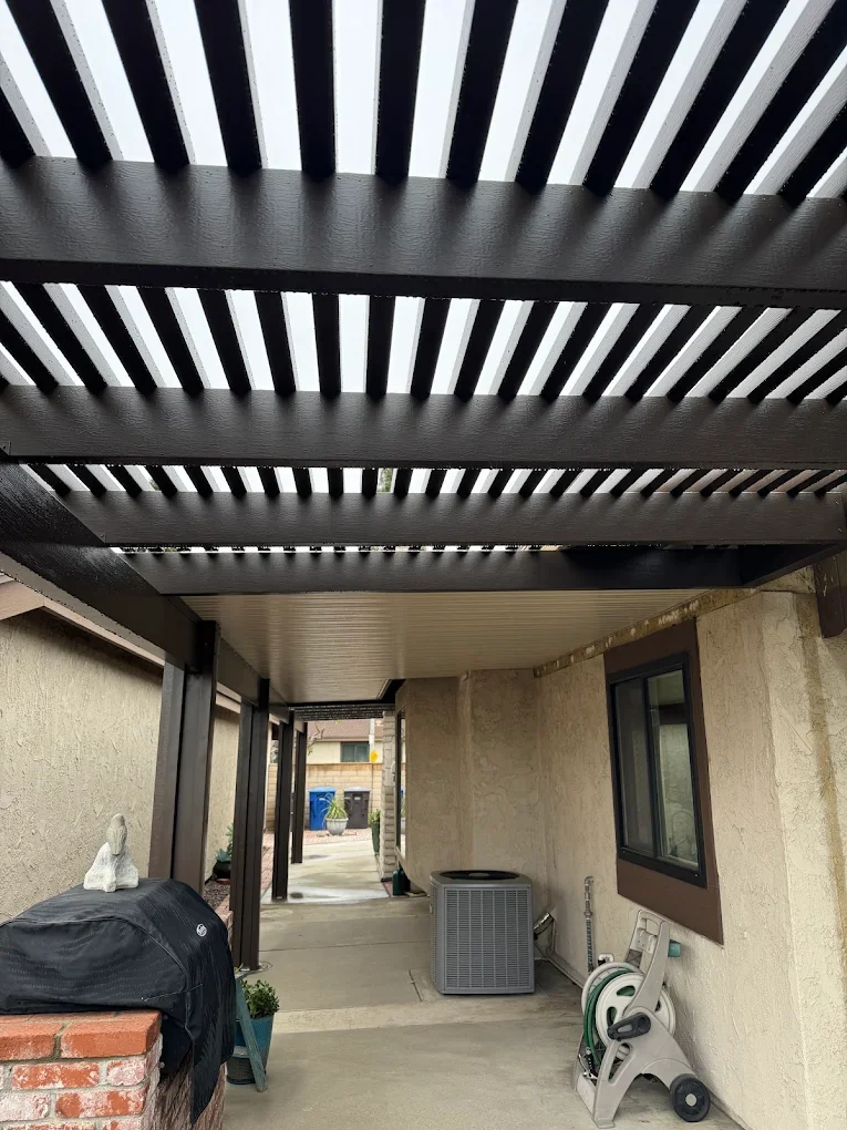 View of a covered patio area with black wooden slats overhead, beige stucco wall, window, and a concrete floor. There is an air conditioning unit, a garden hose reel, and a covered grill with a small decorative white rabbit statue on top.