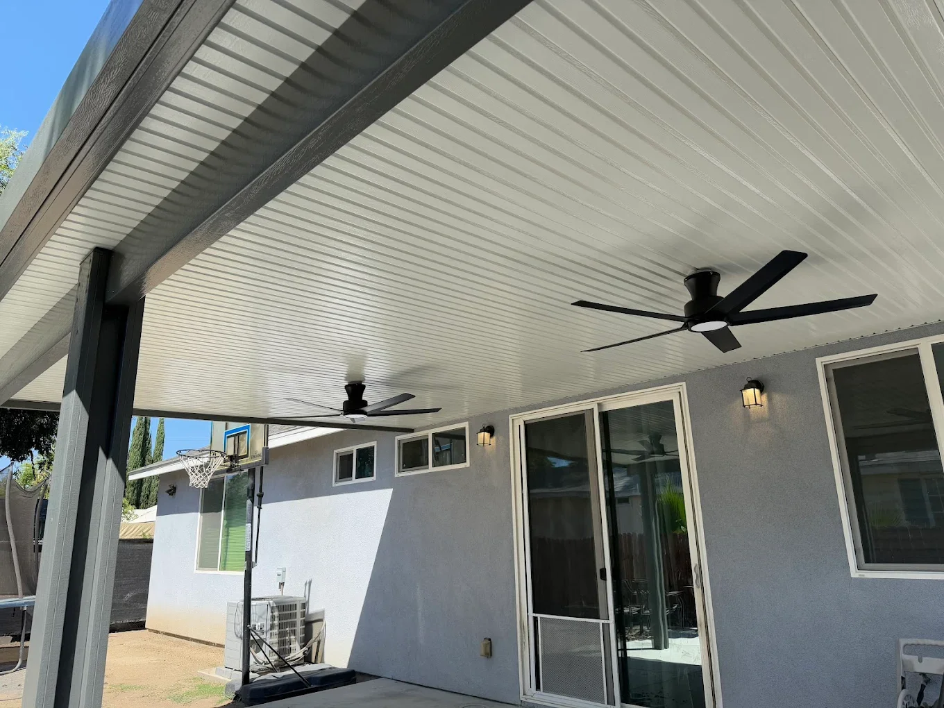 Covered patio with ceiling fans, outdoor lighting, and a sliding glass door, with a basketball hoop and an air conditioning unit visible outside.