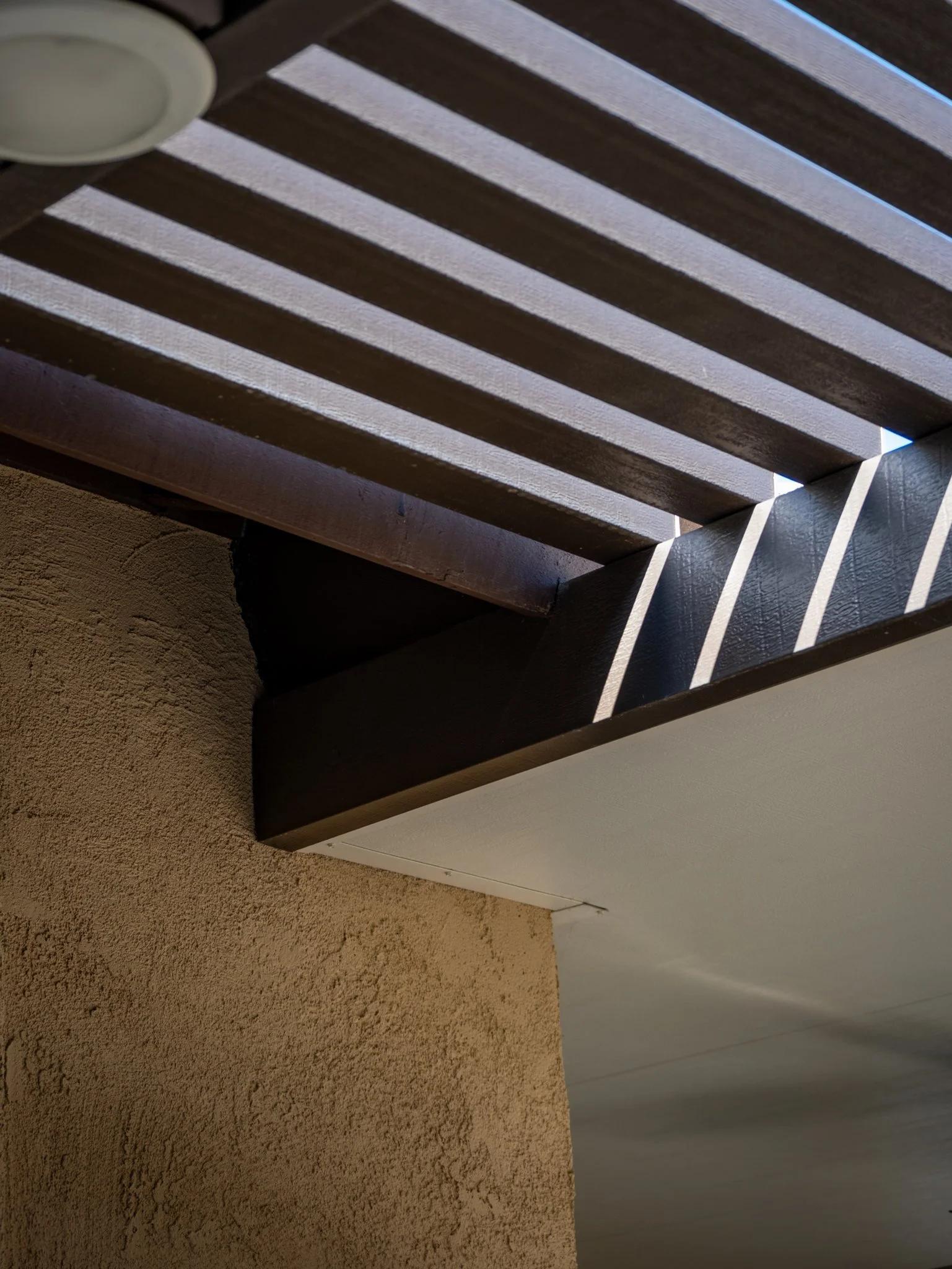 Close-up of a wooden ceiling with sunlight streaming through the gaps, casting shadows on a textured beige wall.