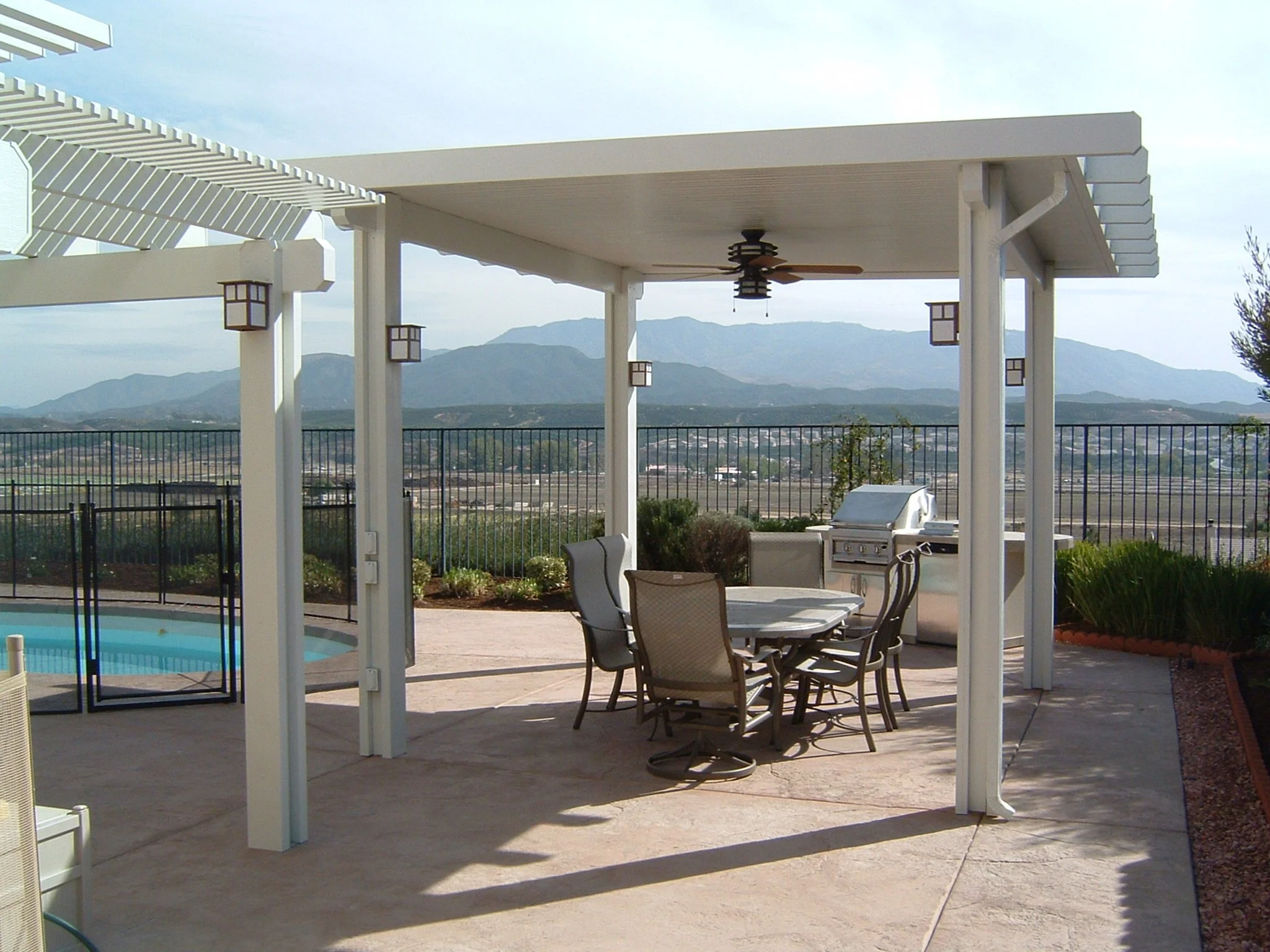 Outdoor patio with a white pergola, a round table surrounded by chairs, a gas grill, and a view of mountains in the distance.