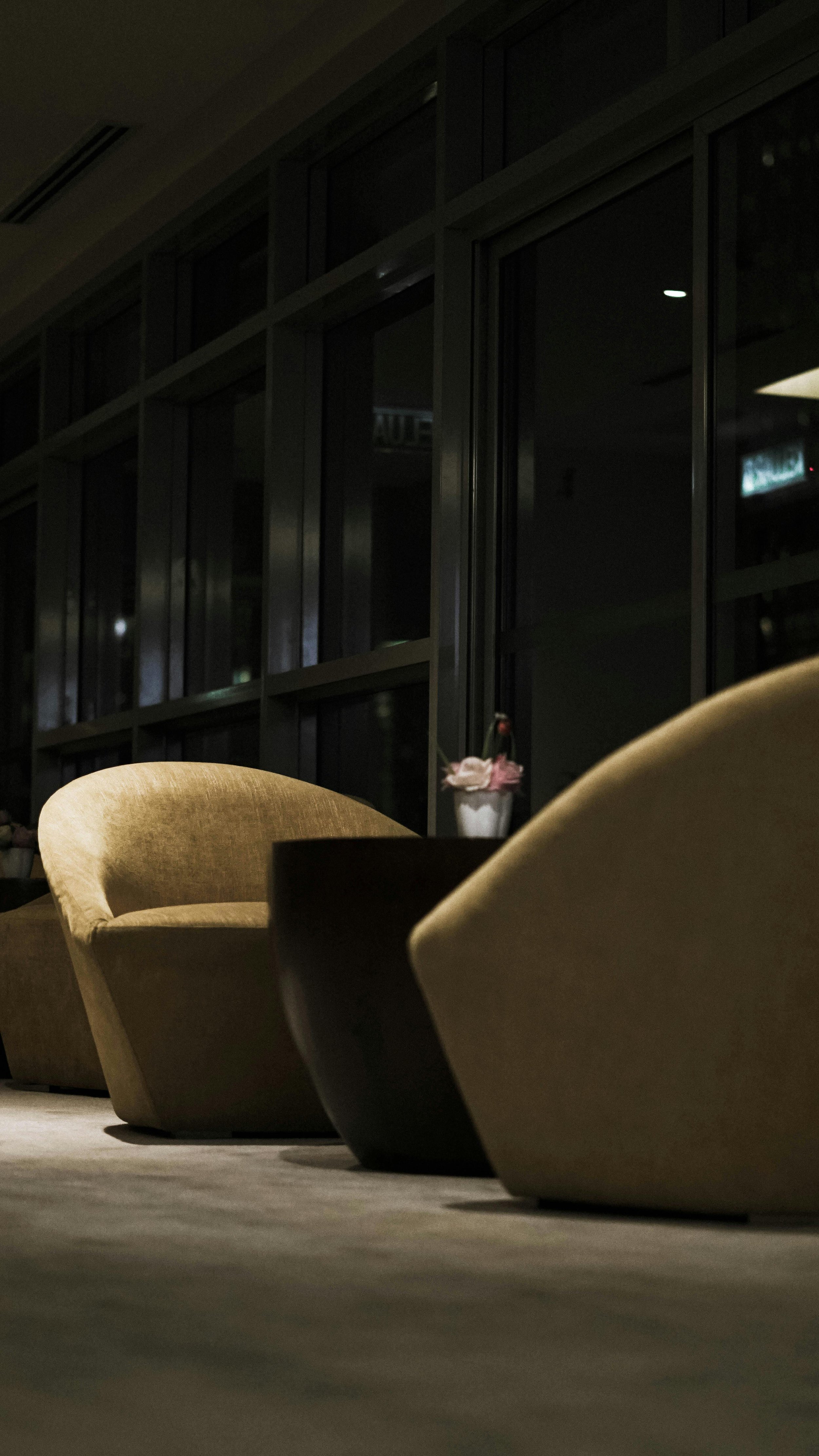 Indoor lounge area at night with beige and dark brown chairs and a low table with a potted plant, against large glass windows.
