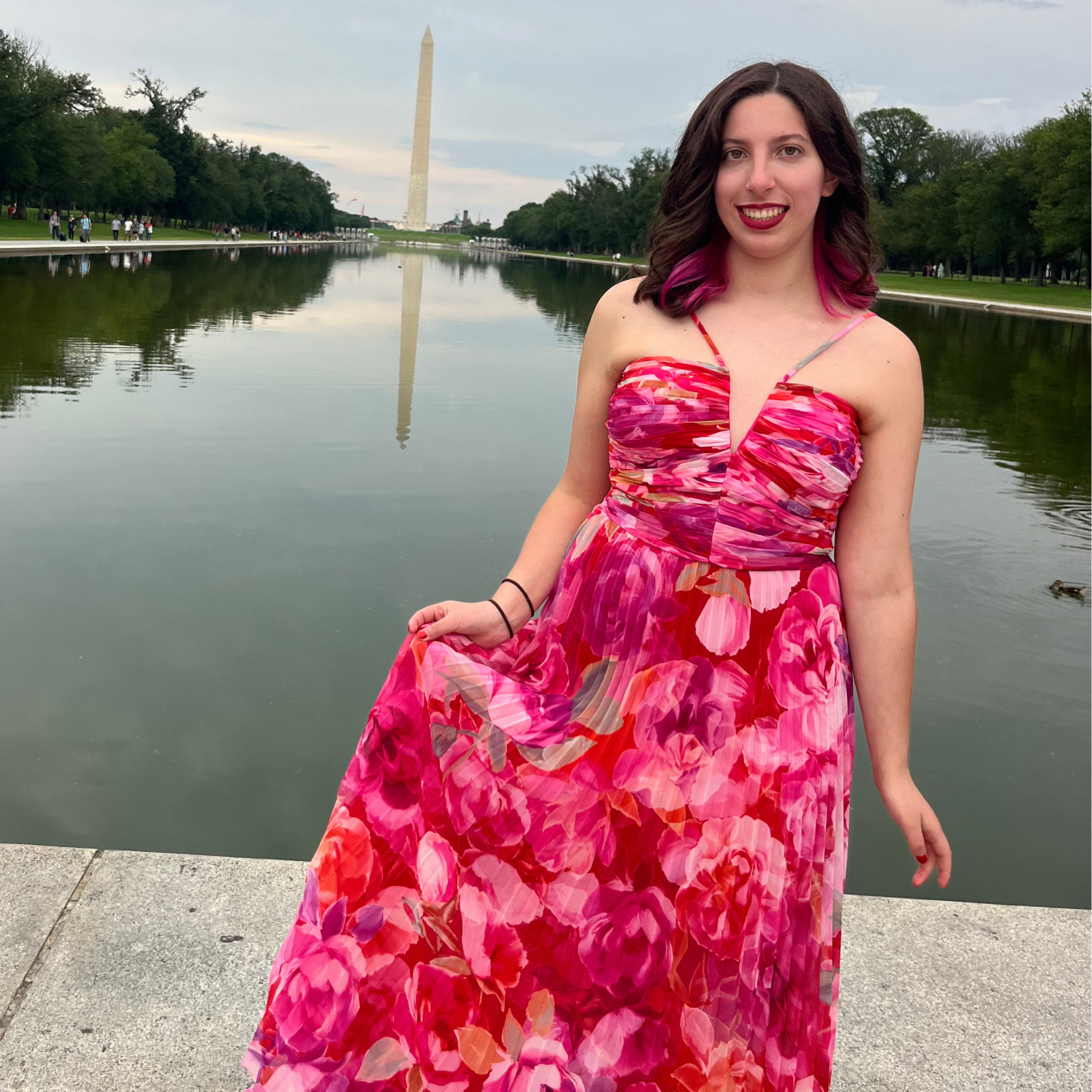A woman in a pink and red floral dress standing by the Reflecting Pool in Washington, D.C., with the Washington Monument reflected in the water behind her.