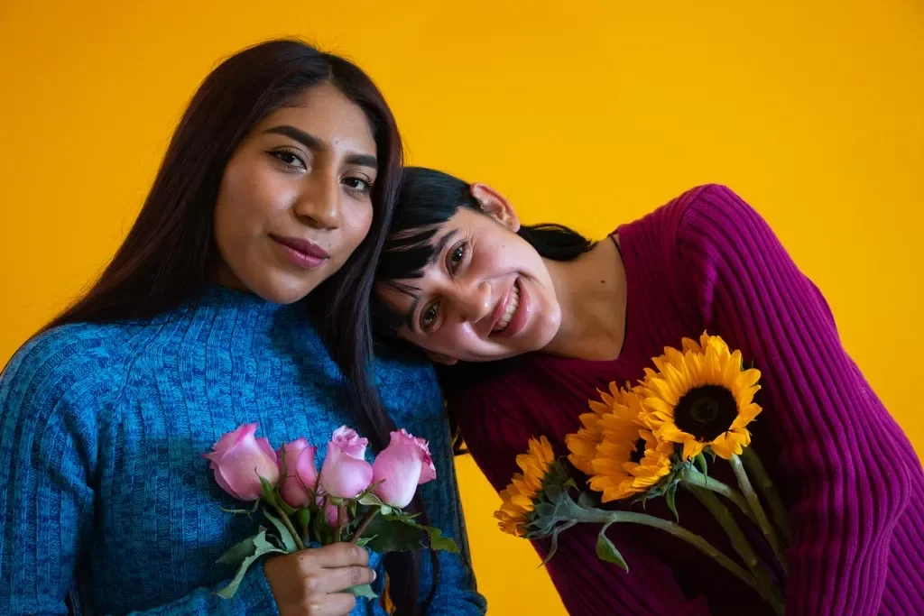 Two women smiling, holding pink roses and yellow sunflowers, against a bright yellow background.