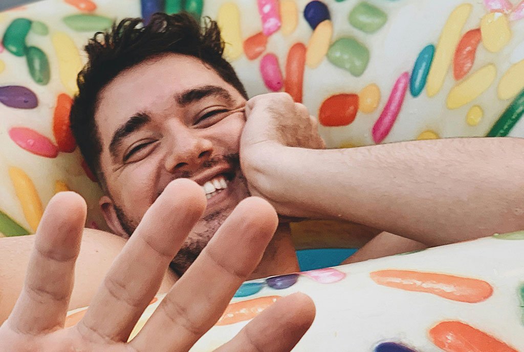 A smiling man lies on a colorful object with sprinkles pattern, making a peace sign with his hand.