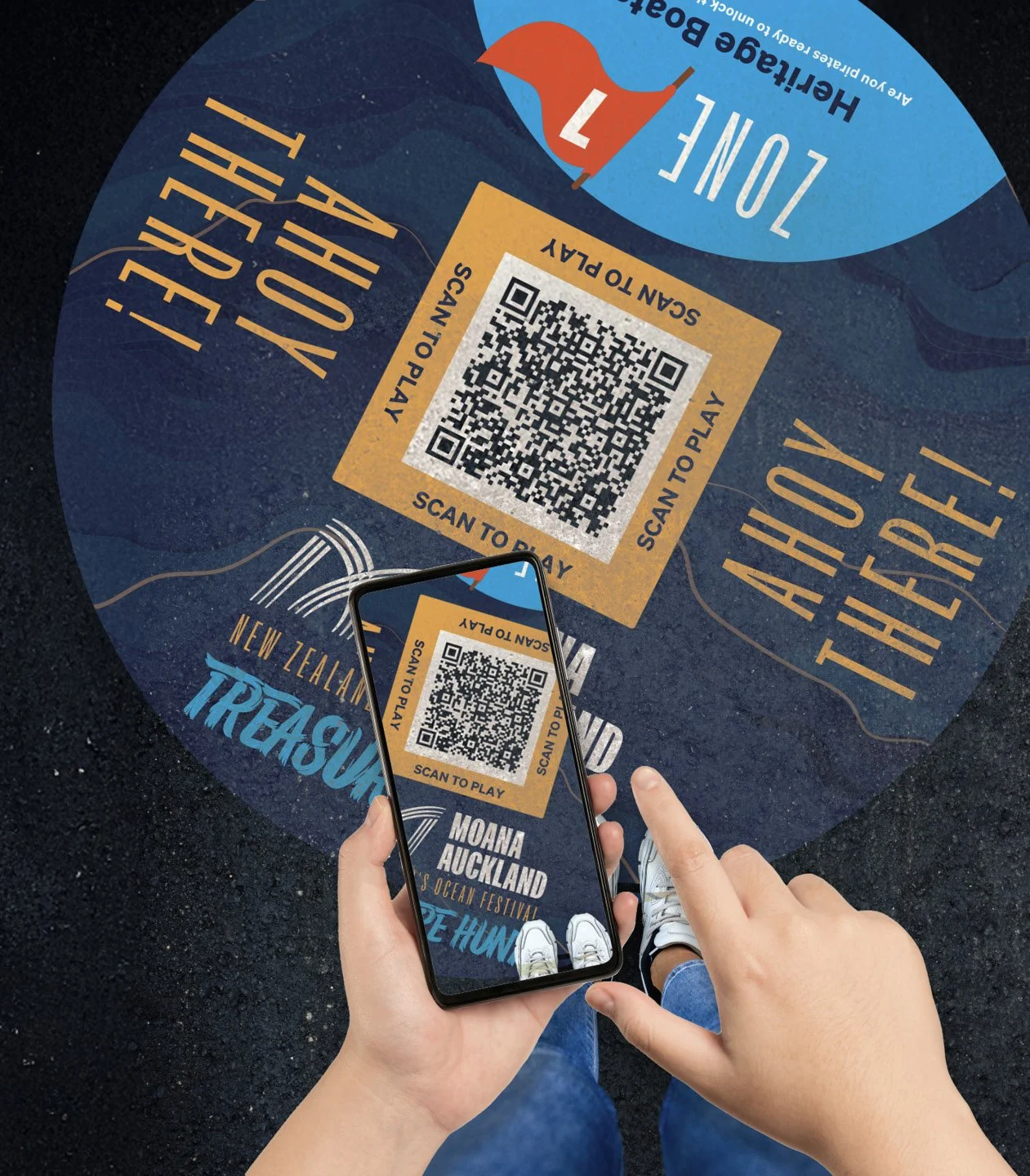 A person holding a smartphone is scanning a QR code on a circular promotional sign for the Moana Auckland Ocean Festival, which has a blue background, a red and blue sailboat illustration, and text promoting a treasure hunt and outdoor activity.
