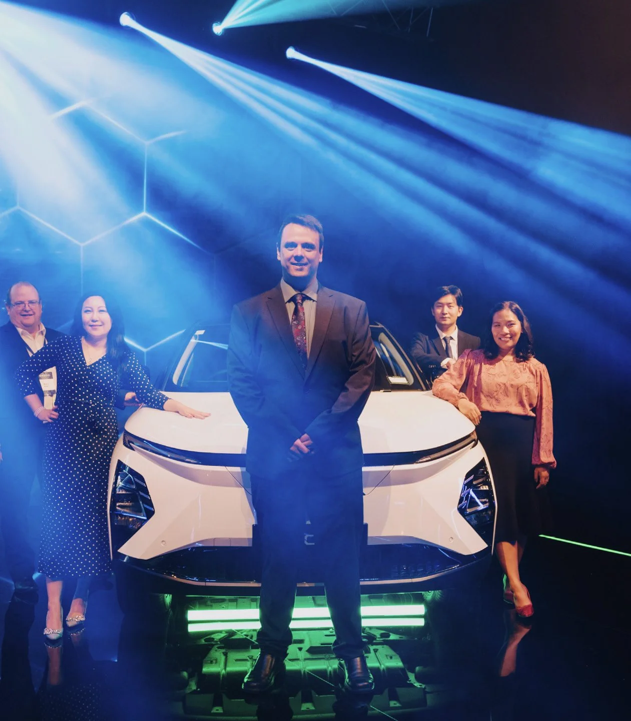 Group of five people standing in front of a white car on a stage with blue lighting and geometric patterns in the background.