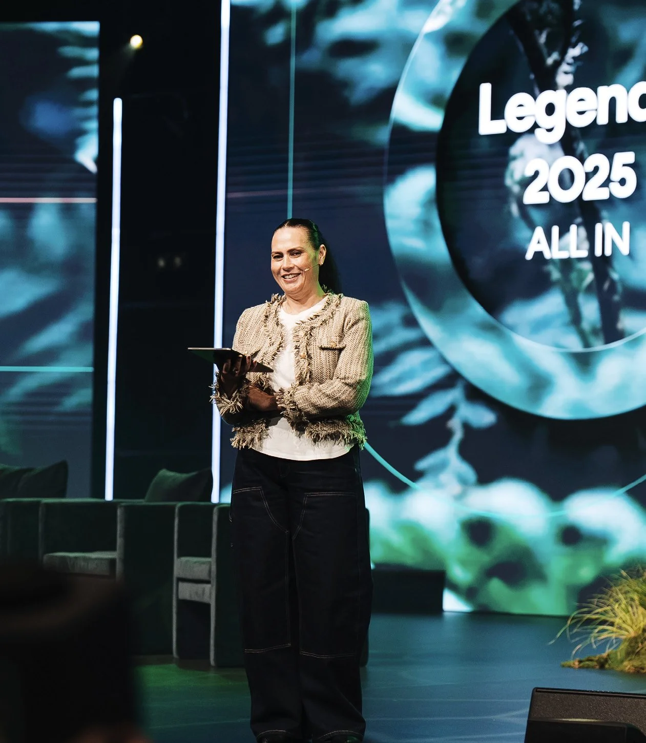 A woman standing on stage, smiling, holding a tablet, with a large digital display behind her that reads 'Legend 2025 ALL IN'.