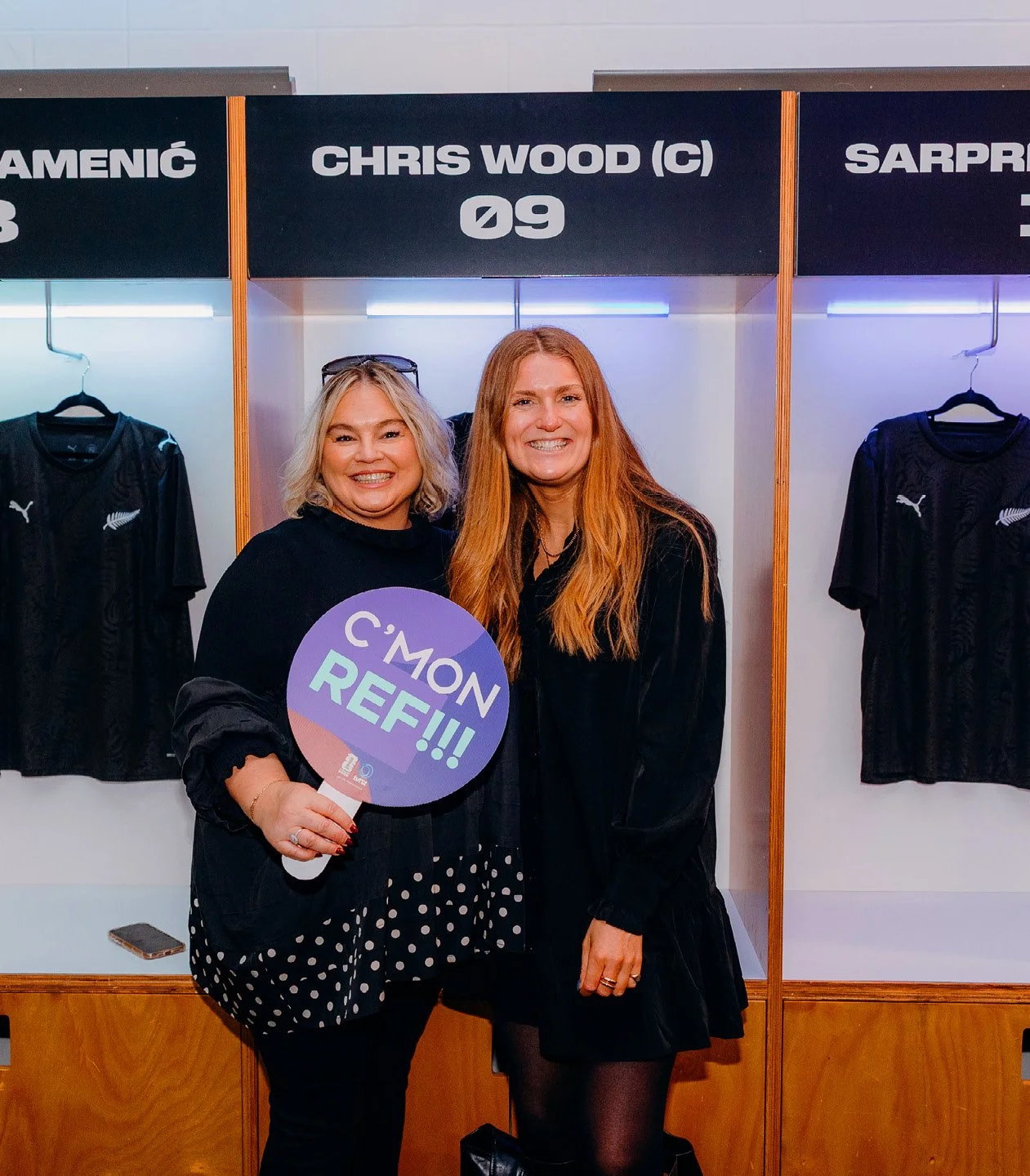 Two women standing in front of a sports locker with black jerseys hanging, one woman holding a sign that says 'C'MON REF!!!' and both smiling at the camera.