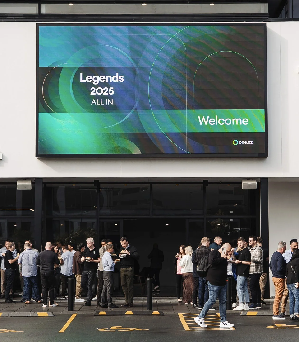 A group of people standing outside a building, waiting to enter, with a large digital screen above them displaying the words 'Legends 2025 ALL IN Welcome' and the logo 'one.nz'.
