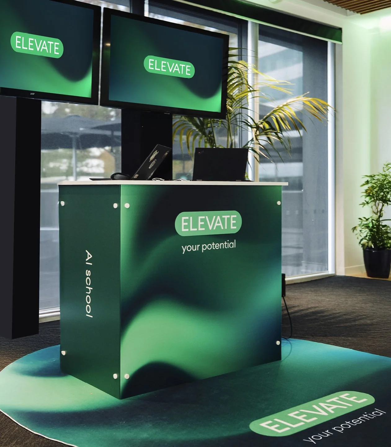 A green and black reception desk with the words "ELEVATE your potential" on the front, located in a modern office with large windows, plants, and two mounted screens displaying the word "ELEVATE".