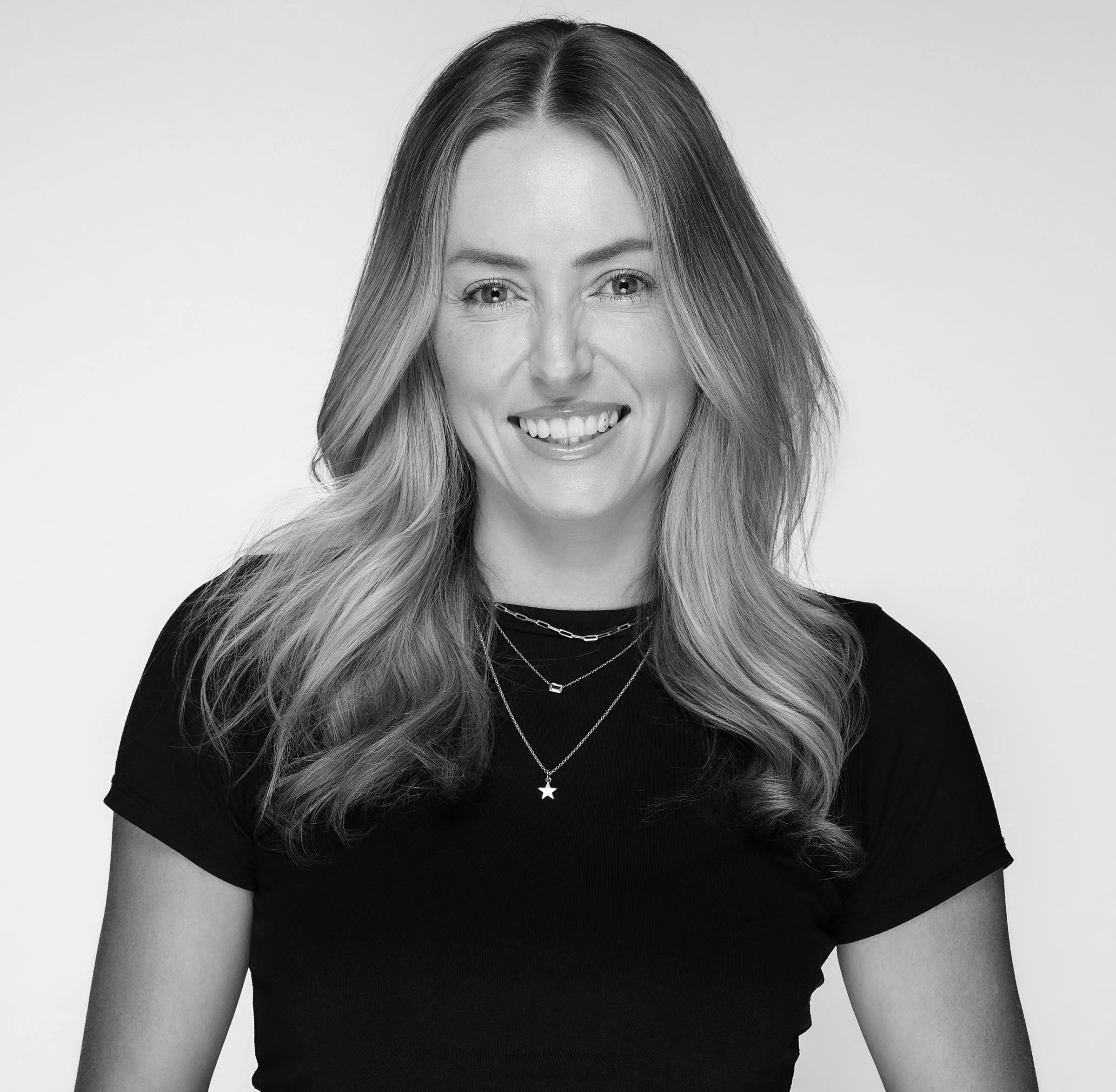 Black and white portrait of a woman with long, wavy hair wearing a sleeveless top, smiling at the camera.