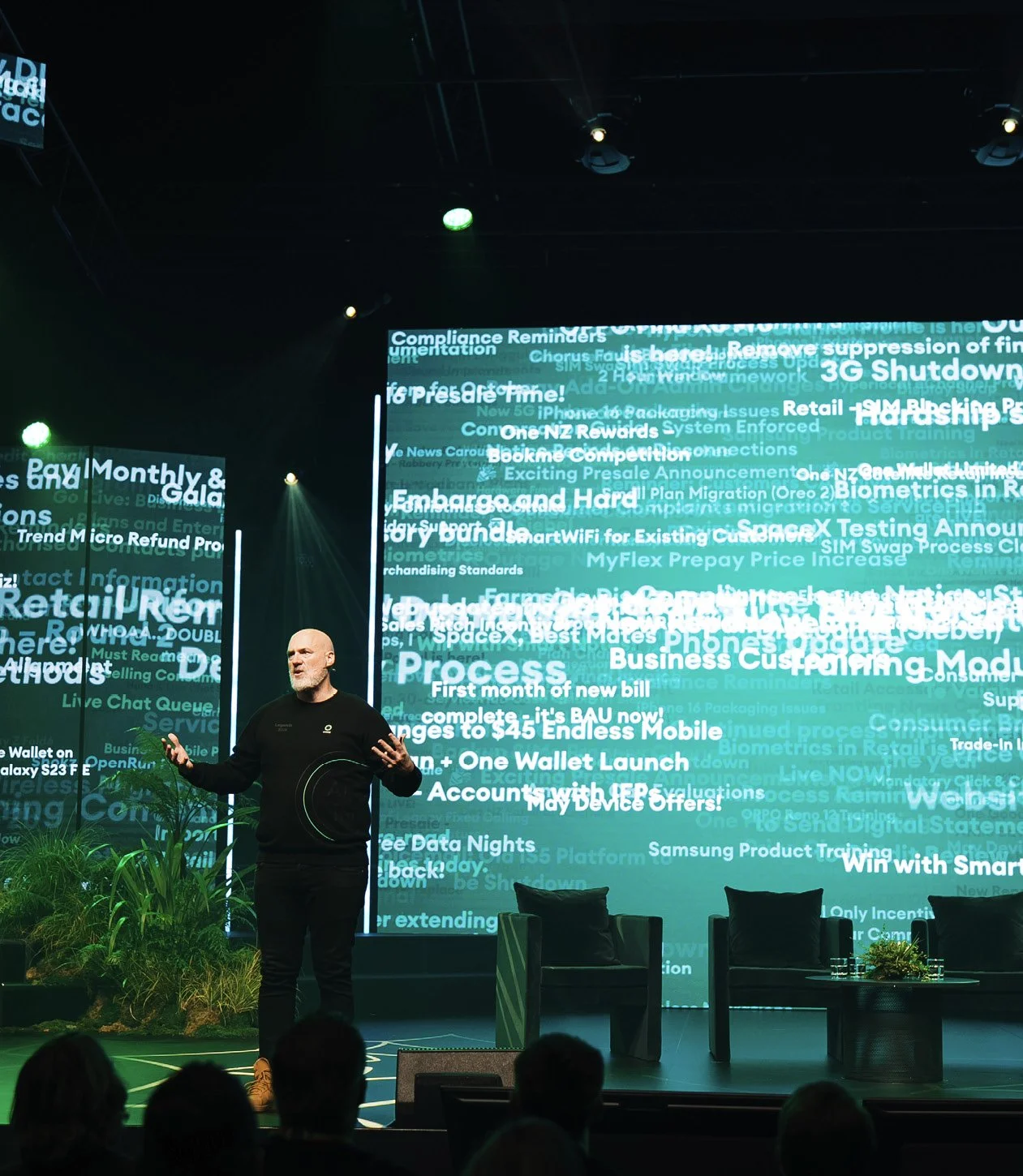 A man with a beard, is standing on stage giving a presentation. Behind him are large screens with various text related to business and technology topics. There are some chairs and plants on the stage, and audience members are visible in the foreground.