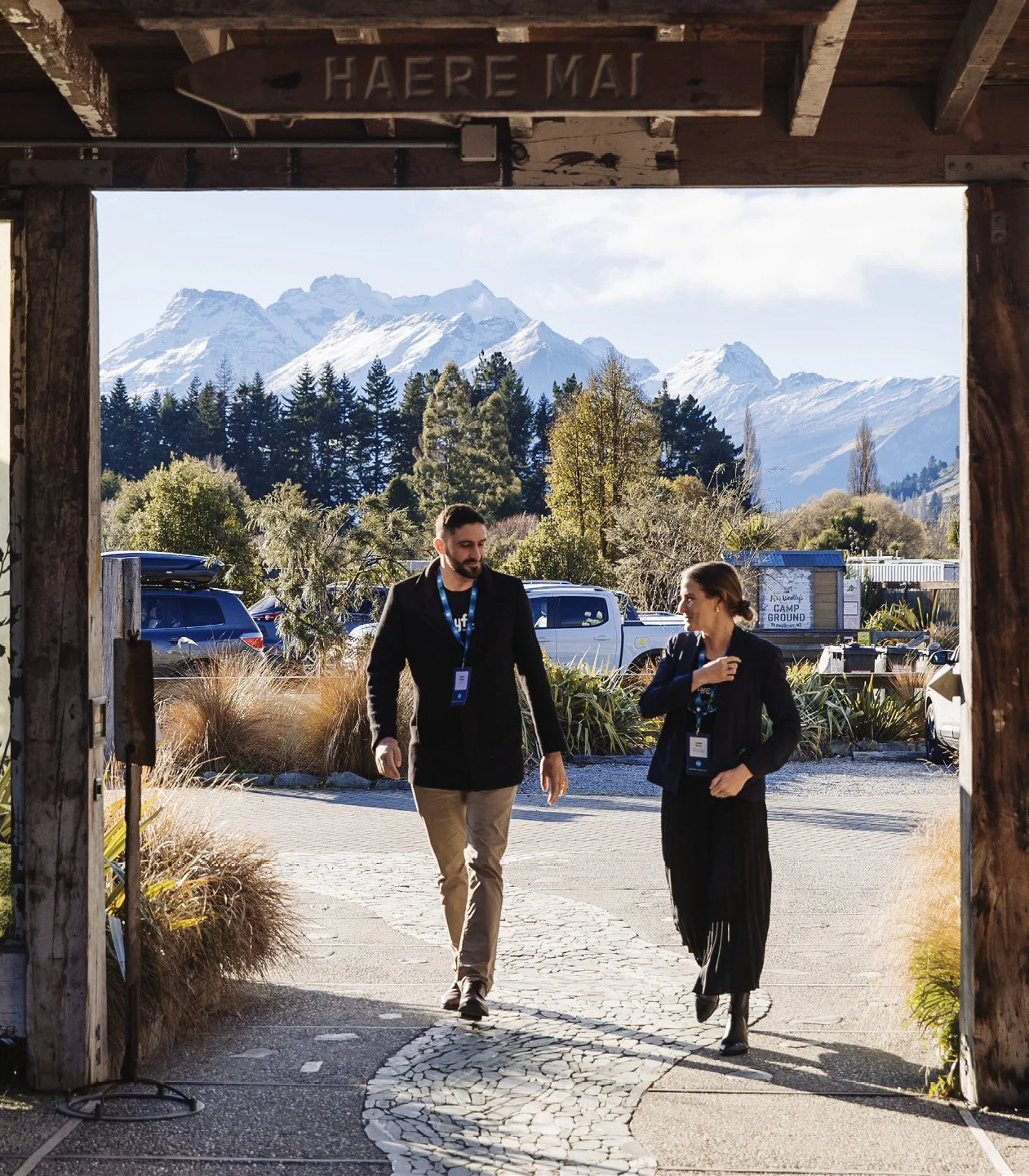 Two people walking under a wooden archway with snow-capped mountains in the background. They are wearing conference badges and outdoors in a scenic area with trees and parked cars.