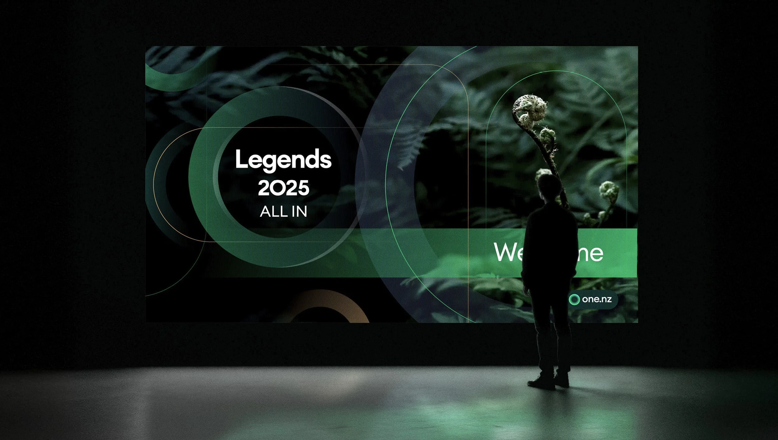 A person standing in front of a large digital screen displaying information about 'Legends 2025 All In' event, with a green-themed graphic background and the text 'Welcome' along with the website 'one.nz'.
