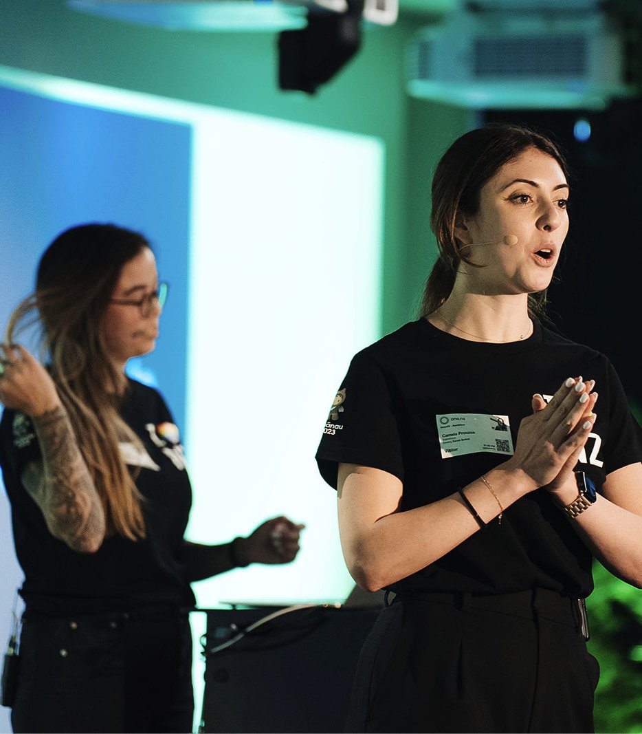Two women are presenting on stage at a conference or event, with one woman in the foreground speaking and the other woman in the background adjusting her hair.