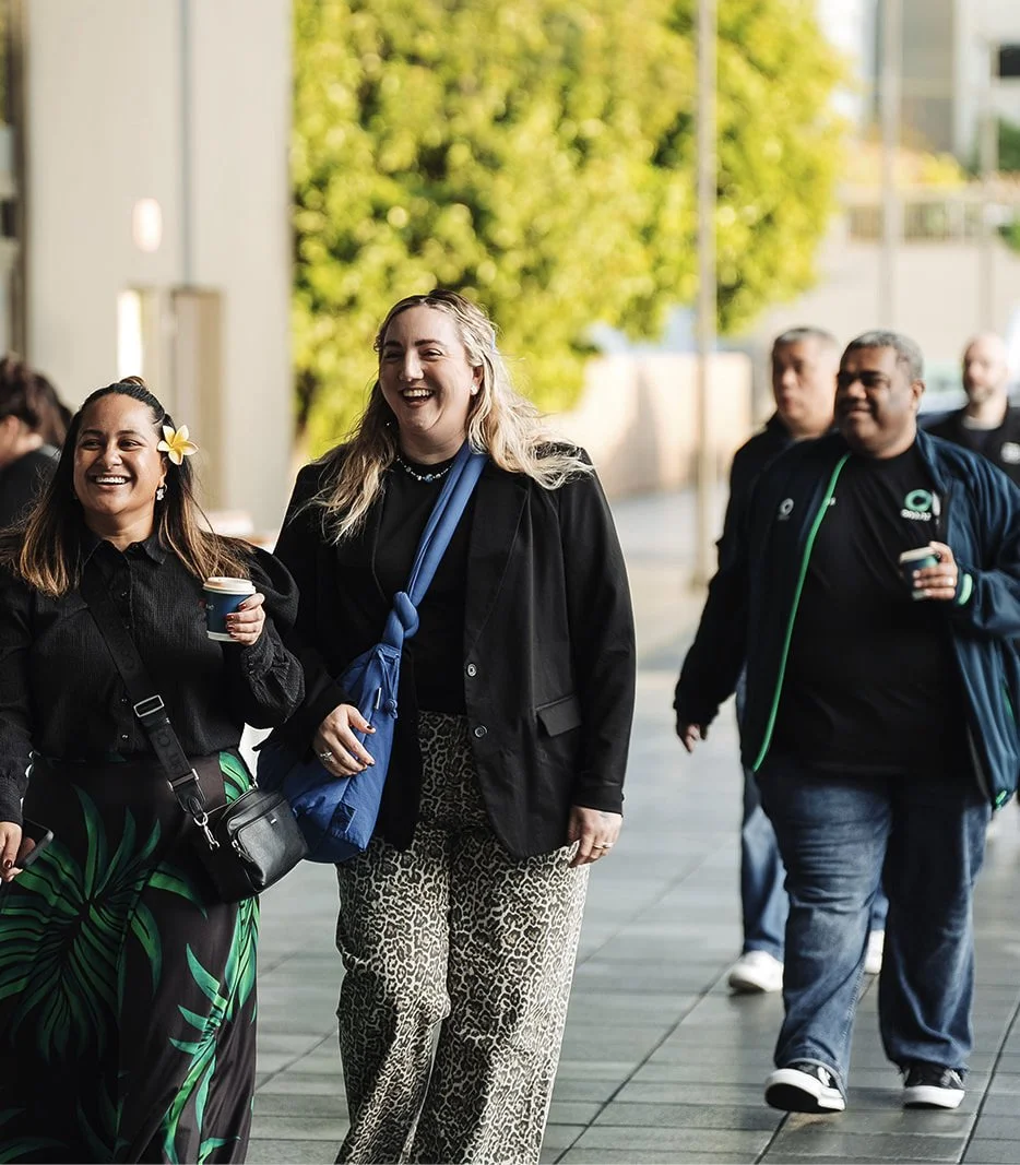 A group of people walking outdoors, smiling and holding drinks, with trees and modern buildings in the background.
