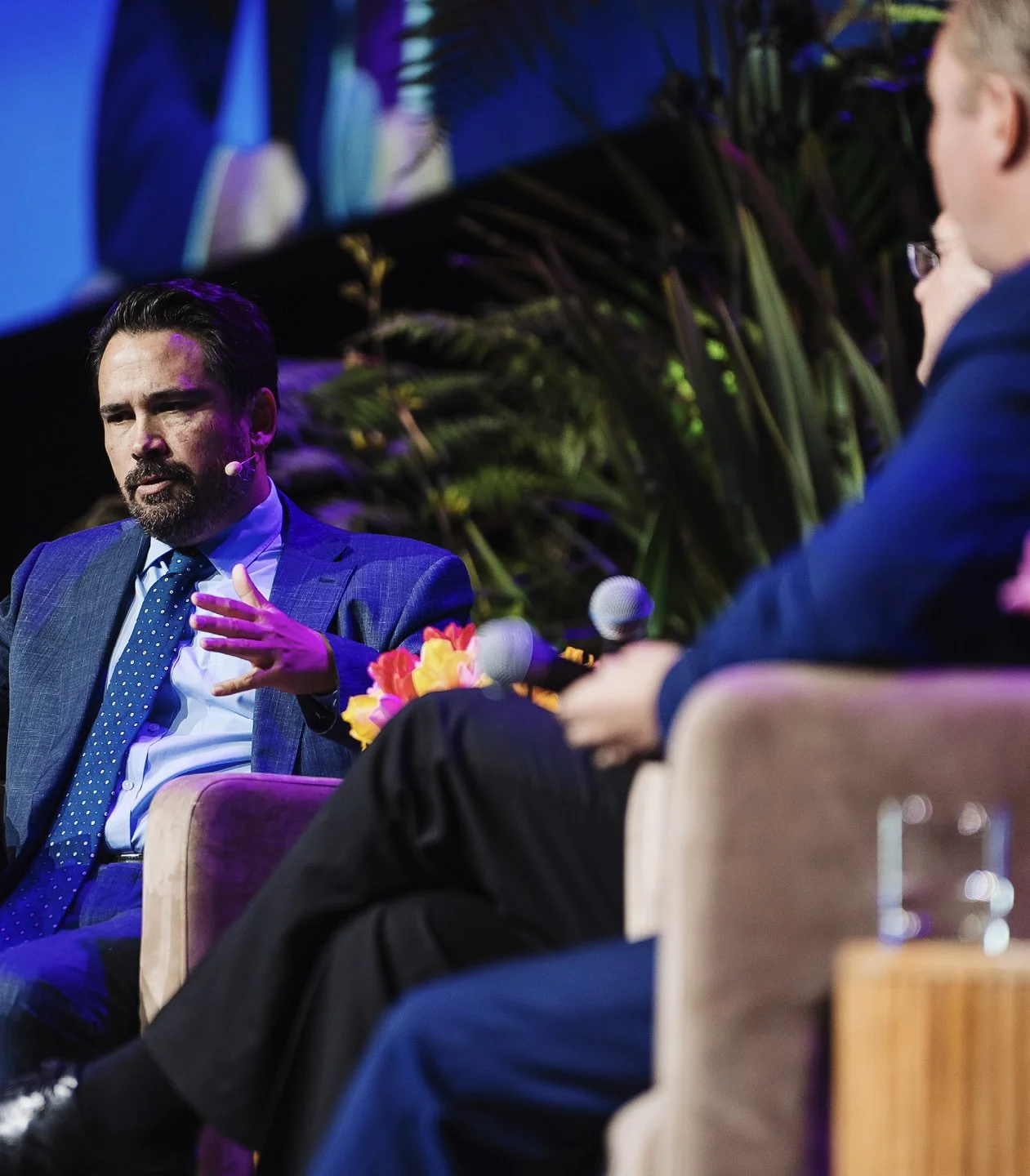 Two men engaged in a discussion on stage at a conference or panel, with one man speaking and gesturing while the other listens, surrounded by plants and decorated with flowers.