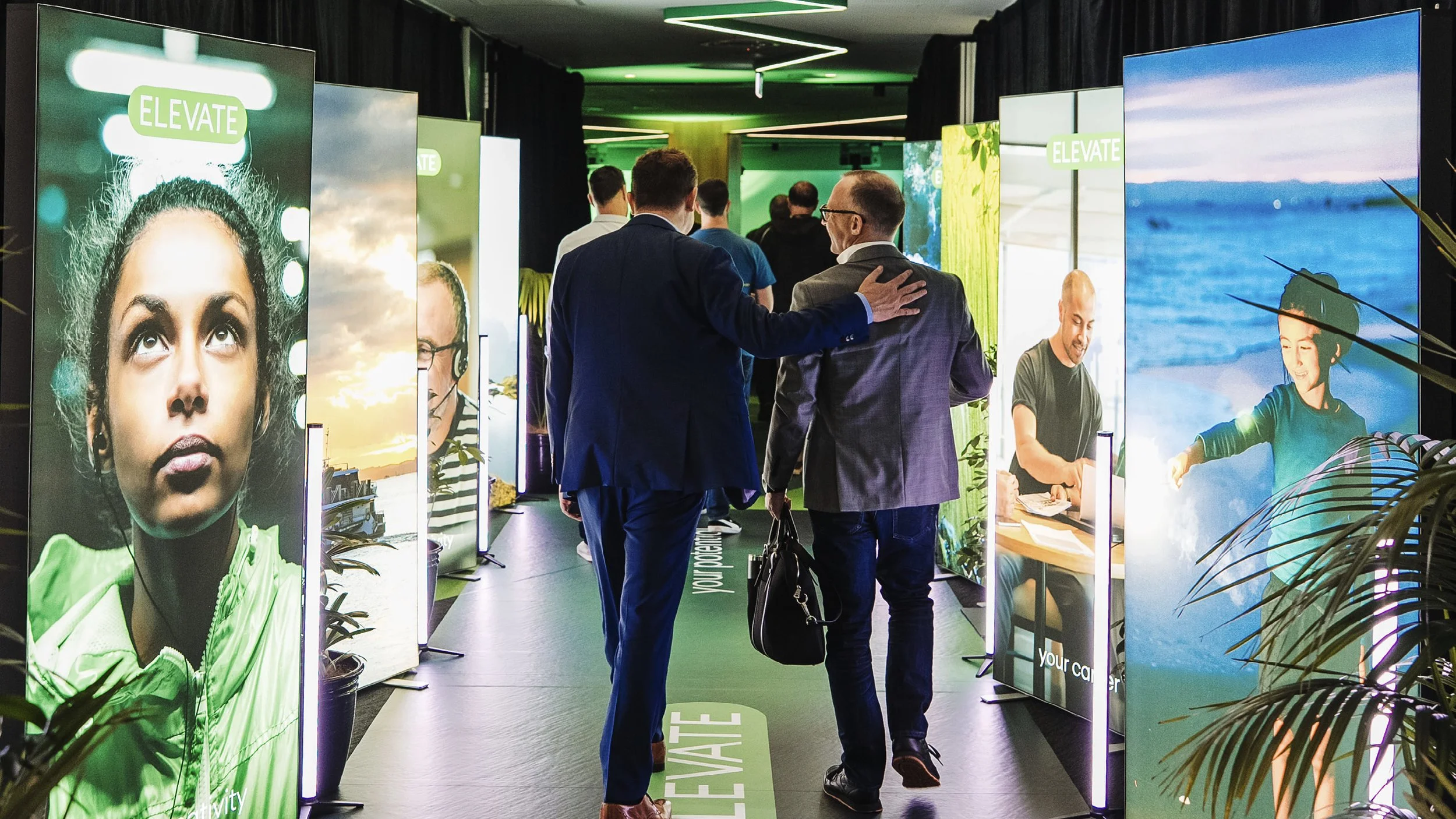 Men in business suits walking through a hallway lined with large illuminated posters featuring people and scenic images at an indoor event or exhibition.