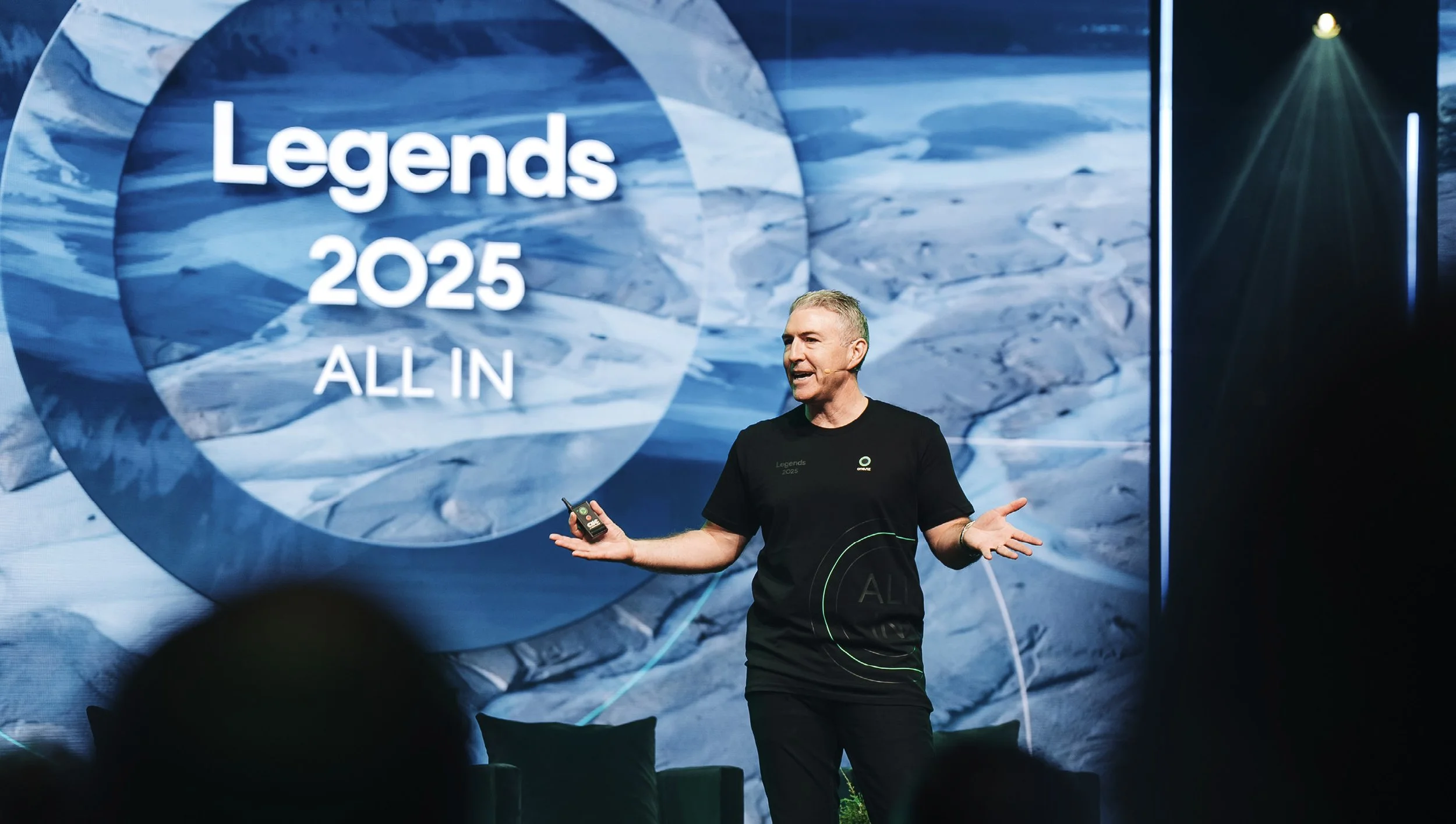 A man talking and gesturing on stage at the Legends 2025 event, with a large screen behind him displaying the event name and slogan.