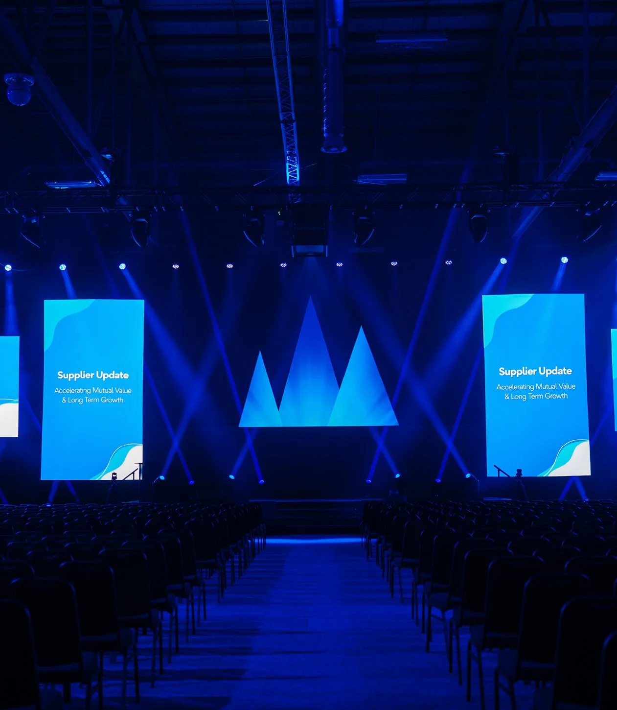 An empty conference or event hall set up with rows of chairs facing a stage with large screens displaying 'Supplier Update' and a blue-themed background.