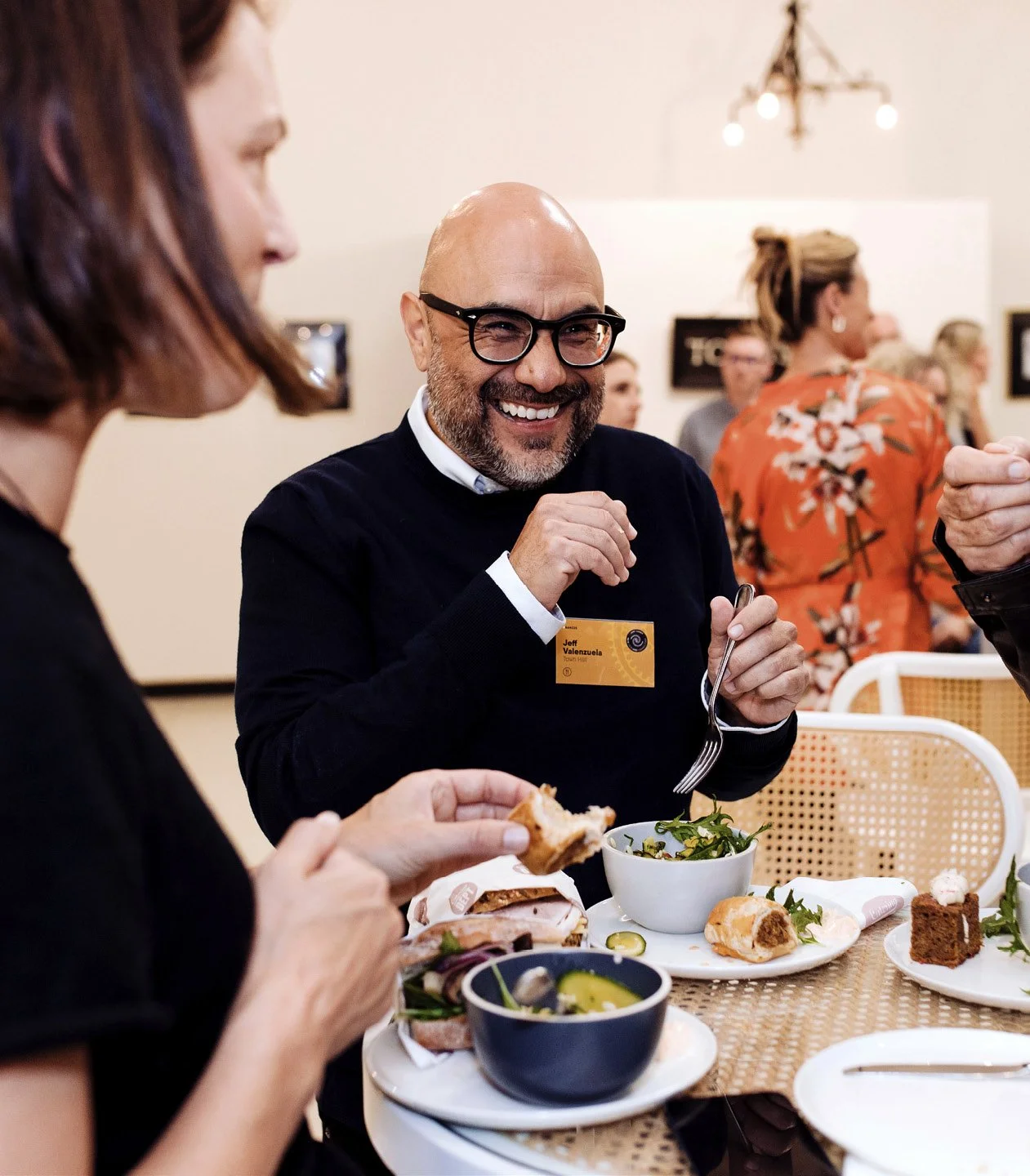 A man with glasses and a beard smiling and eating salad at a restaurant table, surrounded by other people