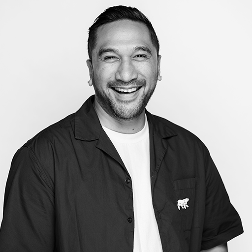 A man with short hair smiling, wearing a black button-up shirt over a white T-shirt, against a plain white background.