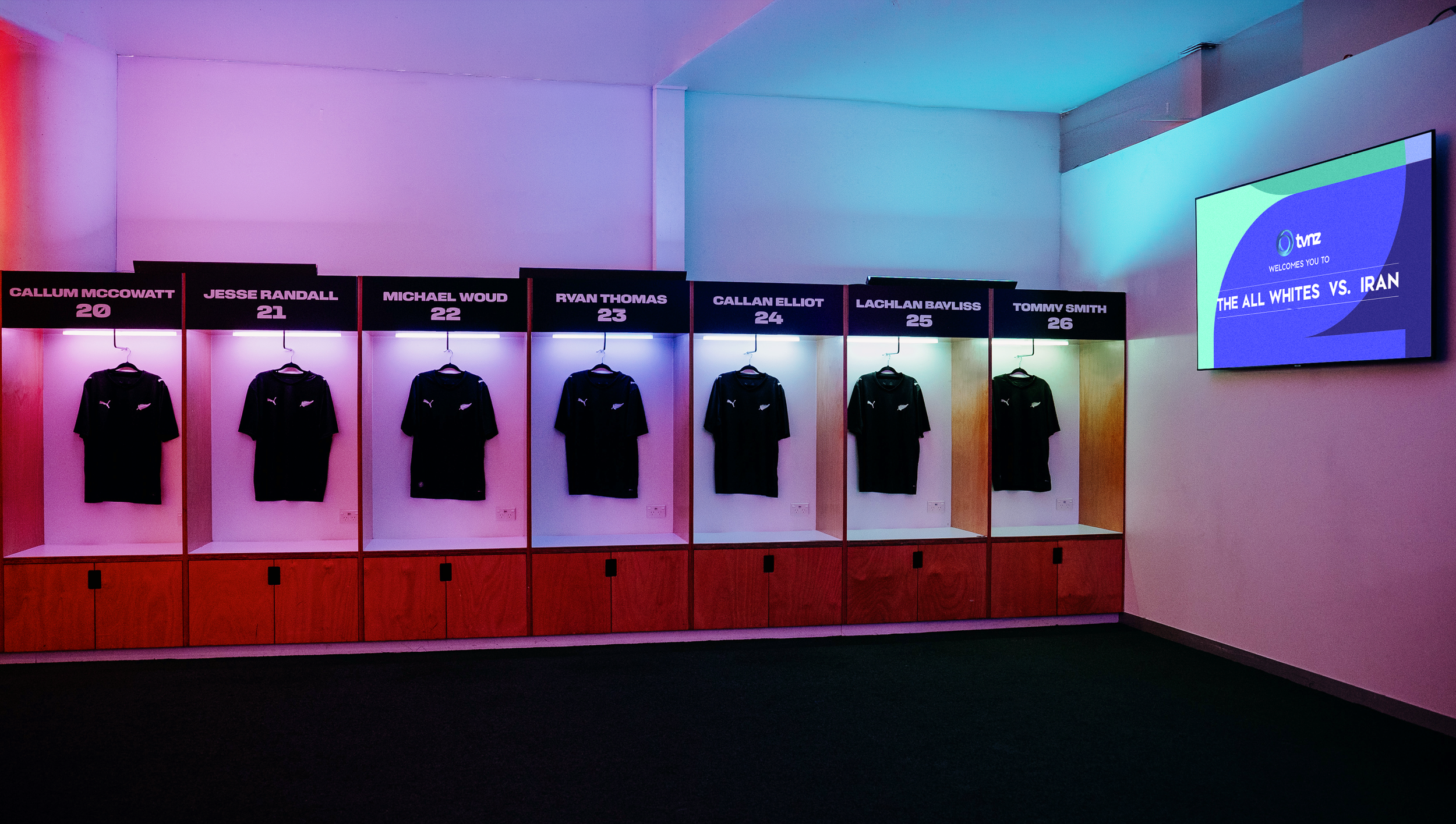 A row of six black jerseys hanging on hooks inside individual wooden cubicles, each with a black sign above labeled with the names Callum McCowatt, Jesse Randall, Michael Woud, Ryan Thomas, Callan Elliot, Lachlan Bayliss, and Tommy Smith, along with their respective jersey numbers 20 through 26. To the right, a large screen displays a message welcoming viewers to 'The All Whites vs. Iran' soccer match under the TNTZ network logo, with a background of colorful gradient.