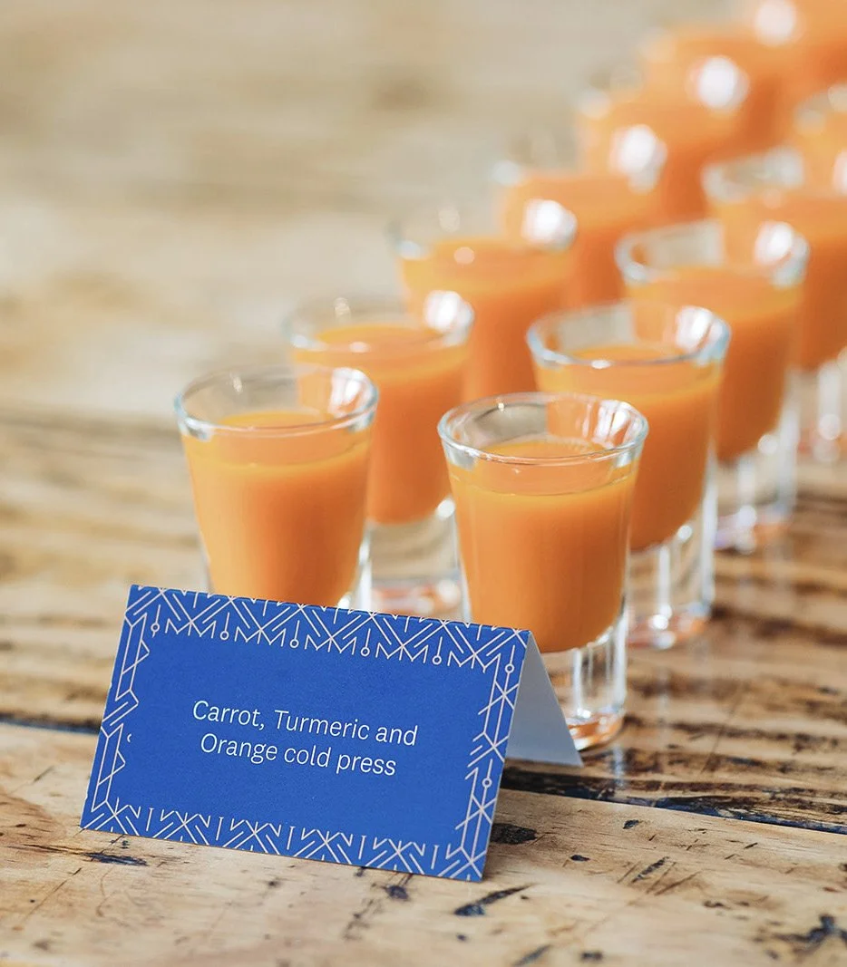 Multiple small glasses containing orange carrot, turmeric, and orange cold press juice on a wooden surface, with a blue sign indicating the juice contents.