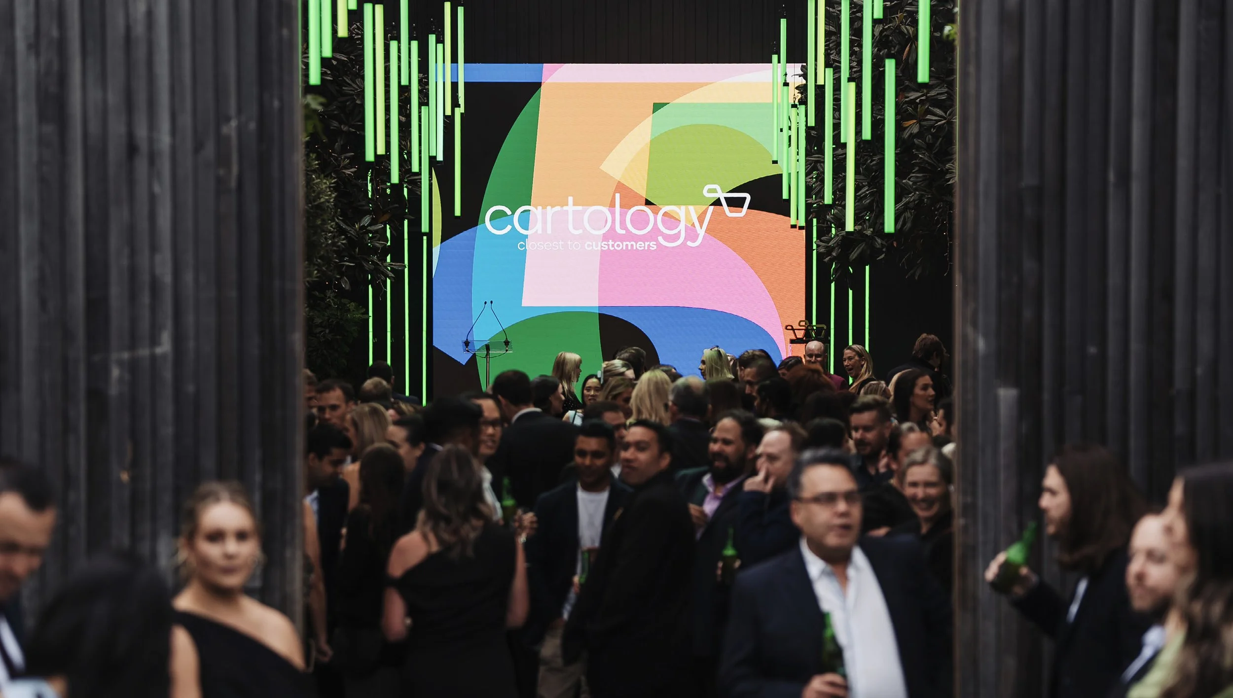 A crowded outdoor event with many people socializing, some holding drinks, in front of a colorful digital screen displaying the logo and name 'Cartology' and the tagline 'closest to customers'. The scene is framed by tall black wooden panels on either side, with greenery visible behind the crowd.