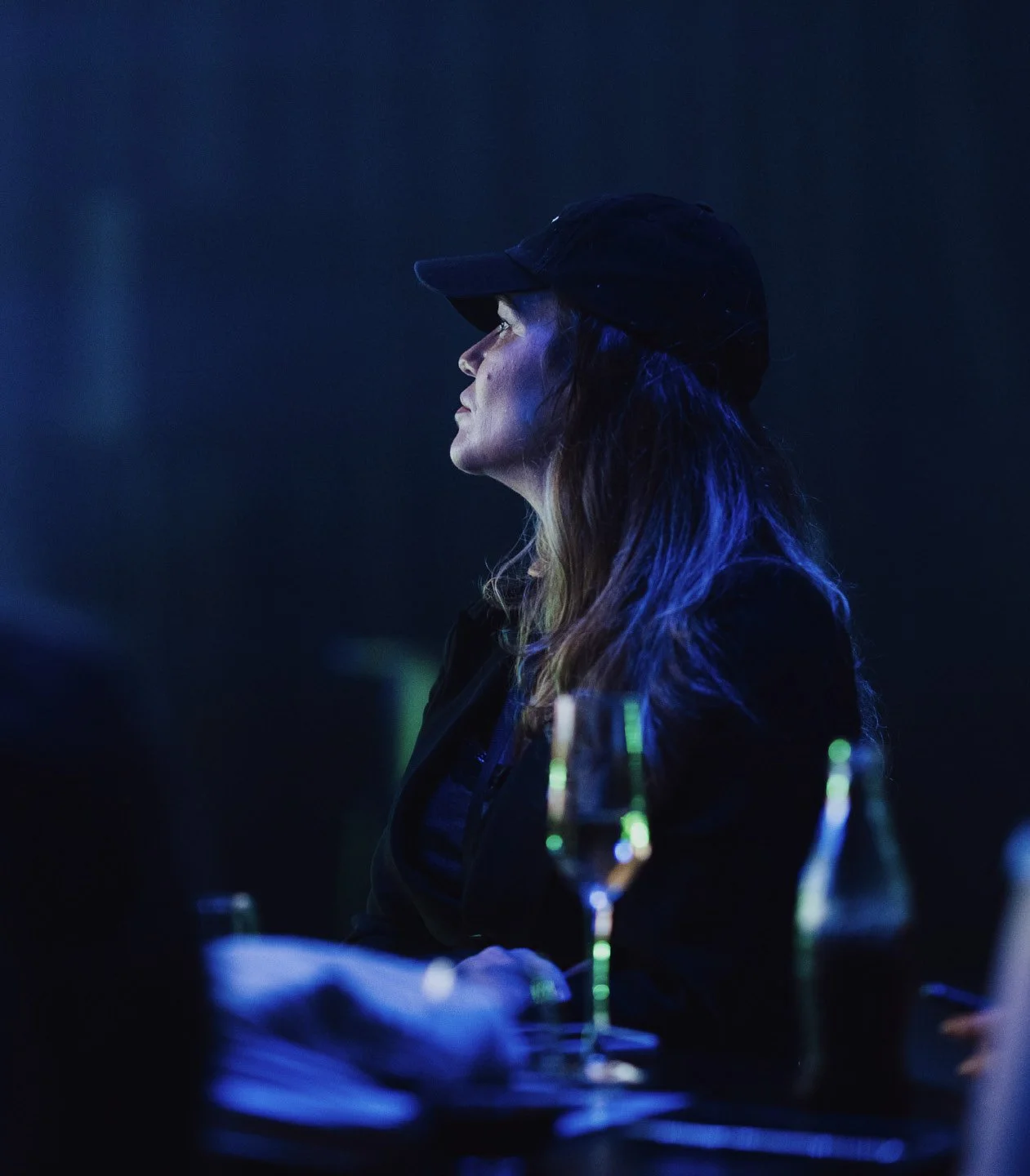 Side profile of a woman wearing a black cap in a dark setting with blue lighting, with a wine glass and a bottle on the table in front of her.
