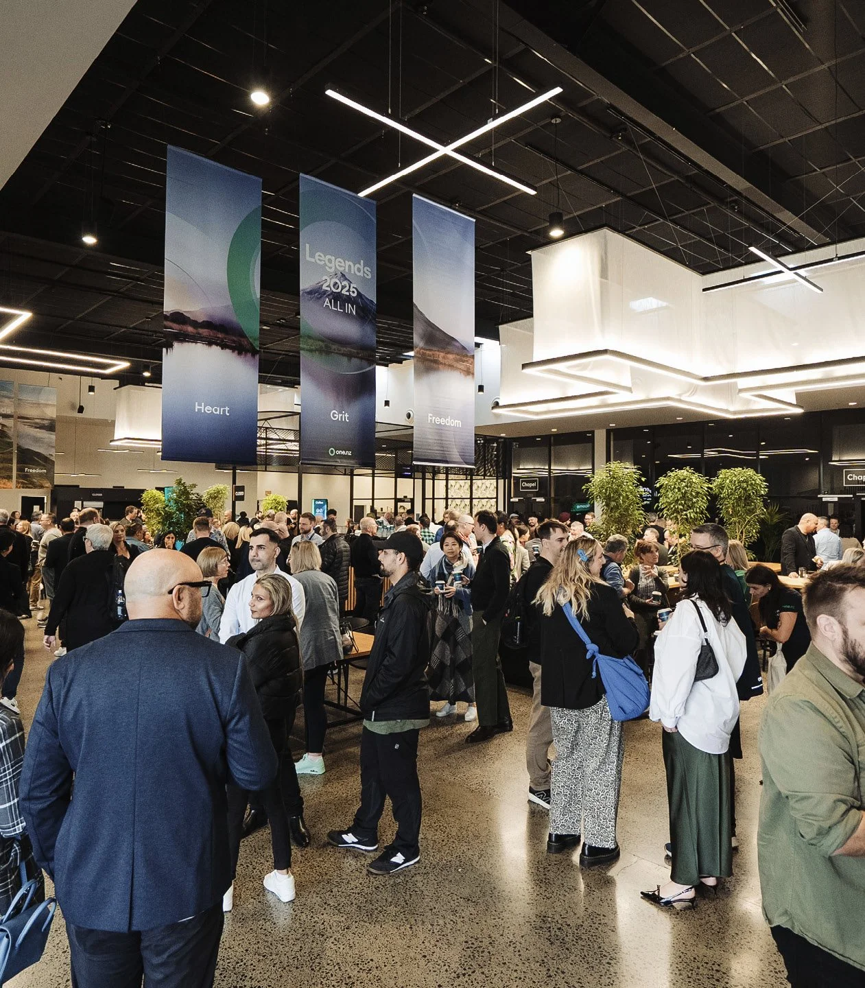 Crowd of people gathered in a modern indoor space with banners hanging from the ceiling displaying words 'Heart,' 'Grit,' and 'Freedom,' and a sign indicating the year 2025.