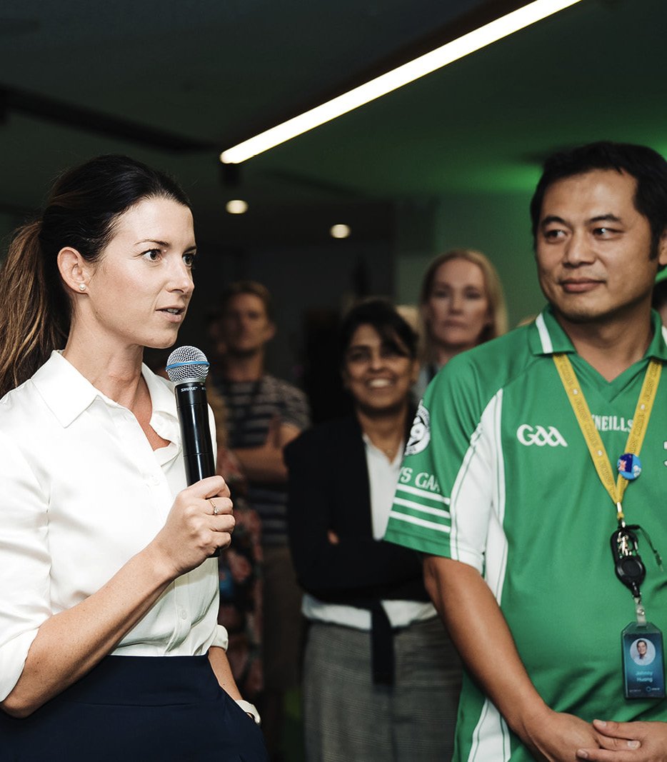 Woman in white blouse holding microphone speaking at an event, with a man in green sports jersey with GAA logo and a lanyard standing nearby, and several women in the background listening.