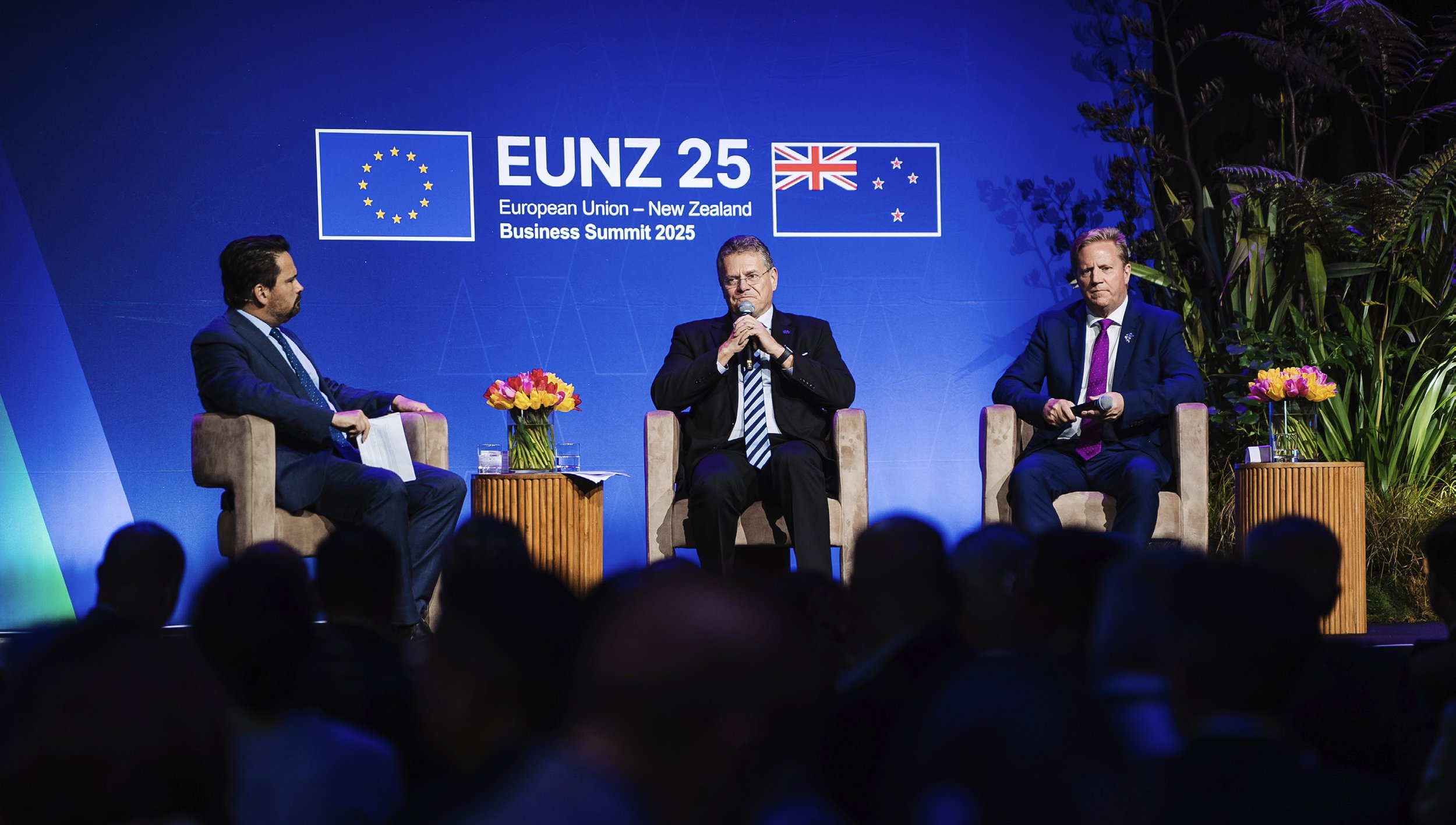 Three men in suits sitting on stage at the European Union-New Zealand Business Summit 2025, with a blue backdrop displaying the event logo and flags of the European Union and New Zealand, decorated with flower arrangements on small tables.